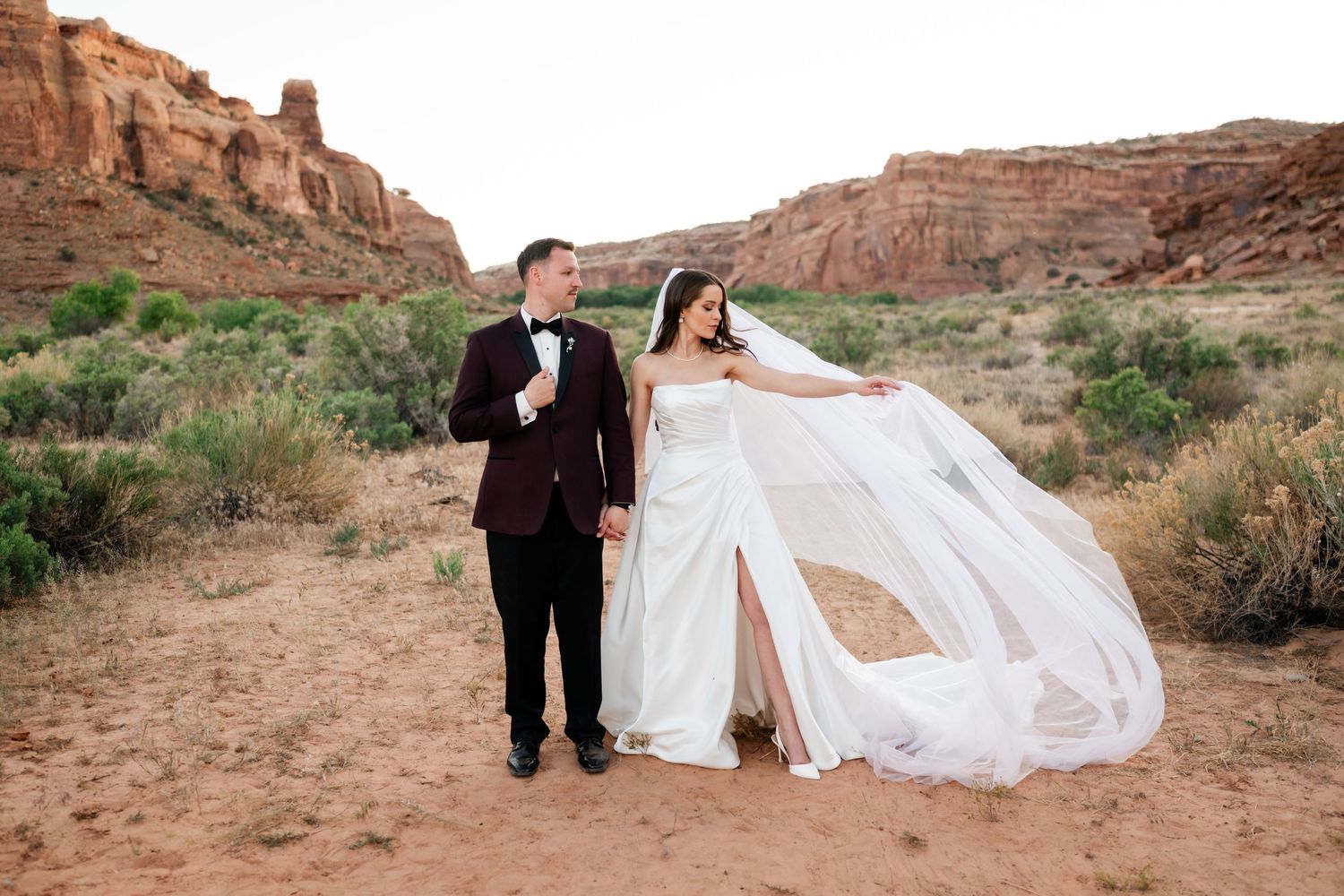 Bride in strapless gown with flowing veil and groom in burgundy tuxedo hold hands in red rock desert canyon.