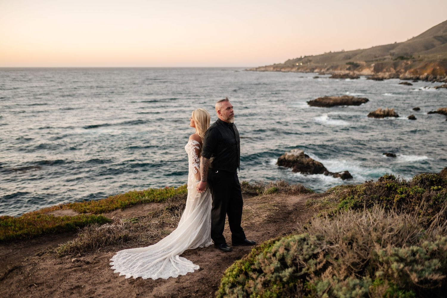 A couple in wedding attire embraces on a rocky coastal cliff during golden hour with waves crashing below.
