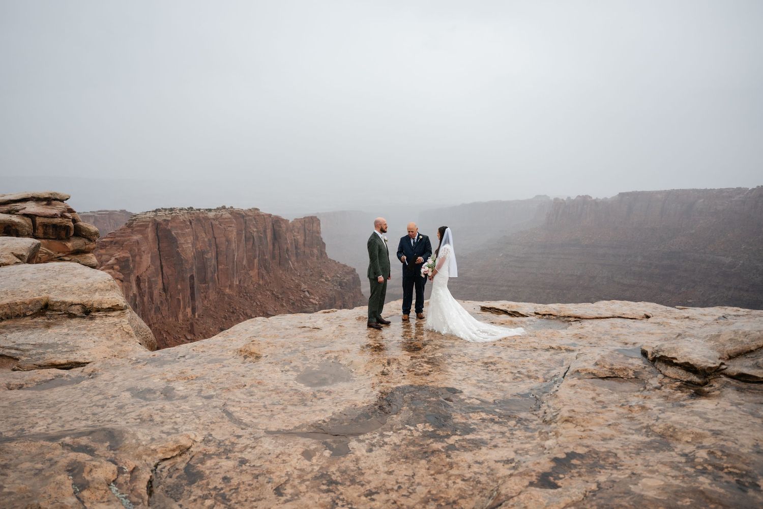Tourists stand at the edge of a vast canyon overlook with dramatic red rock cliffs stretching into the misty distance.