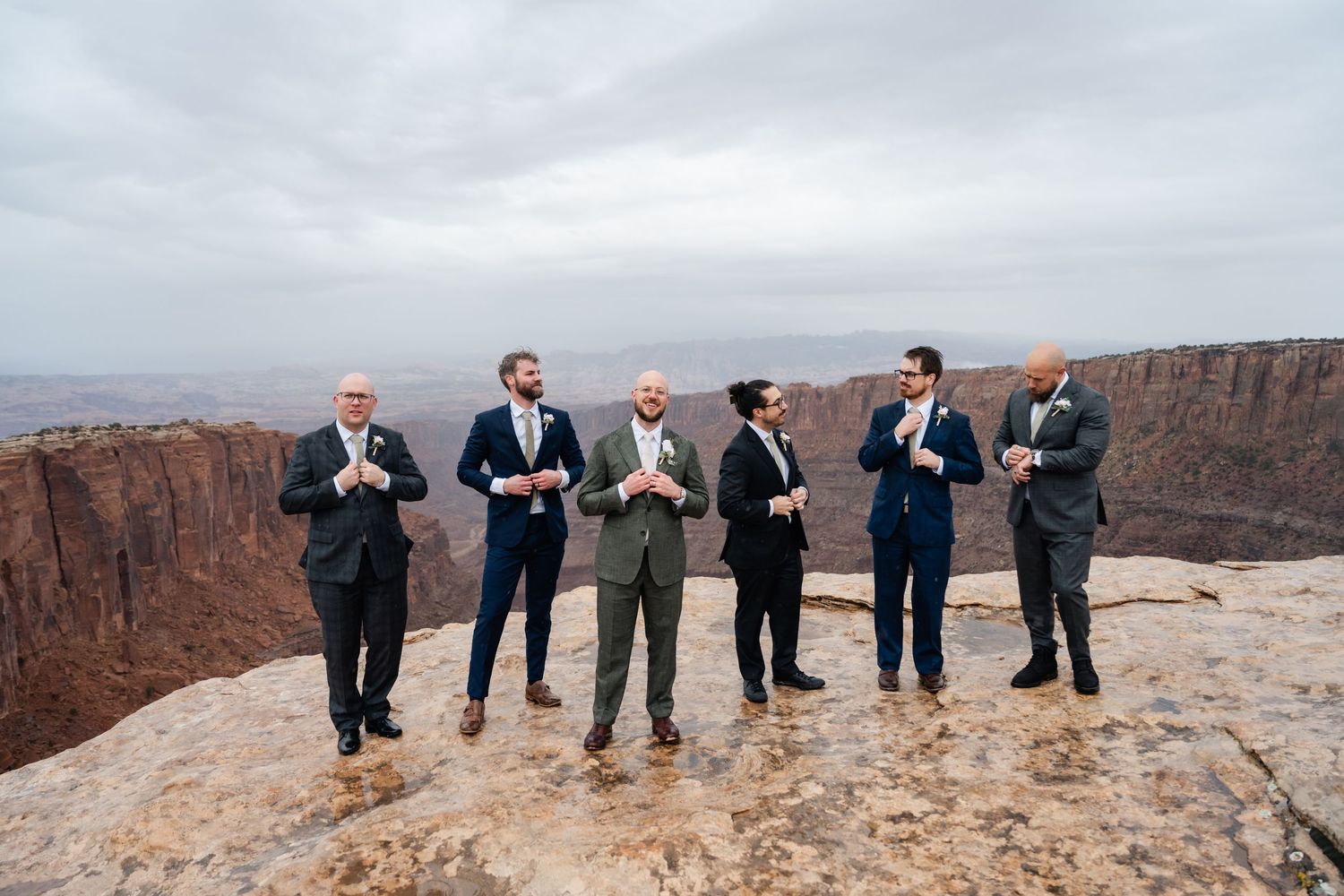 Group in formal attire poses along edge of scenic desert canyon overlook with cloudy sky.