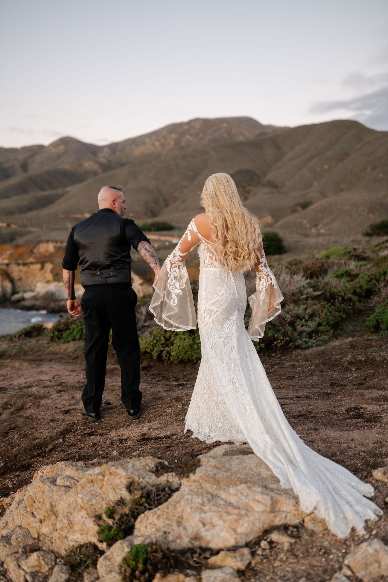 Couple in wedding attire walking along rugged coastal cliffs at sunset.