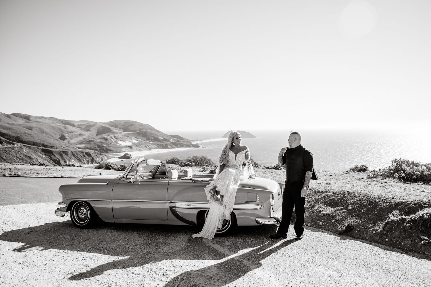 Black and white photo of couple with vintage car on coastal highway.