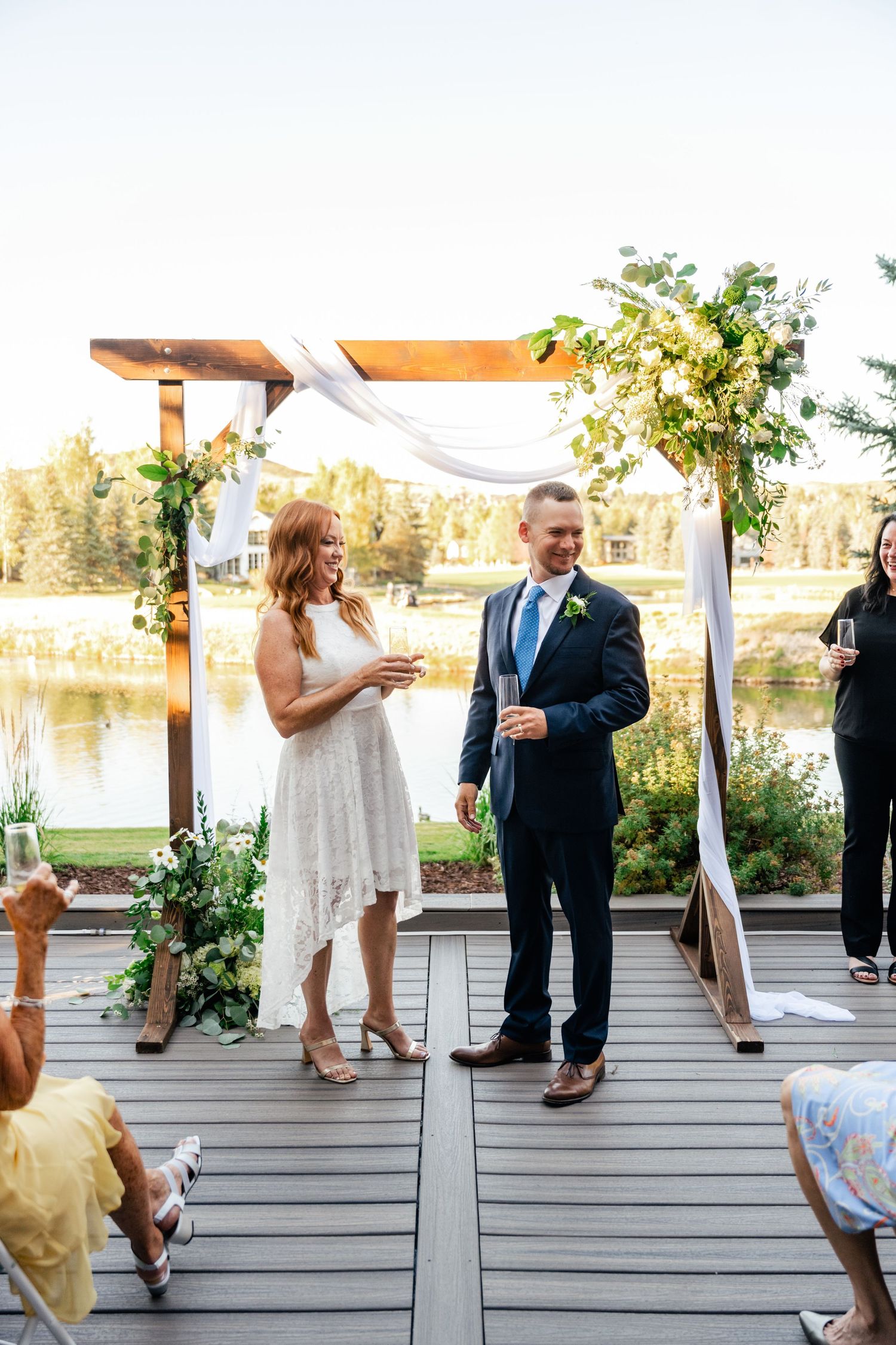 Wedding ceremony unfolds on rustic wooden deck with lake and forest views at sunset.