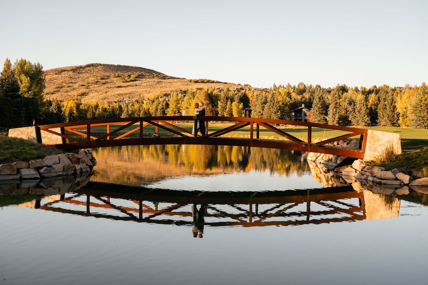 A wooden footbridge reflects perfectly in calm lake waters during golden hour with autumn trees and hills in background.