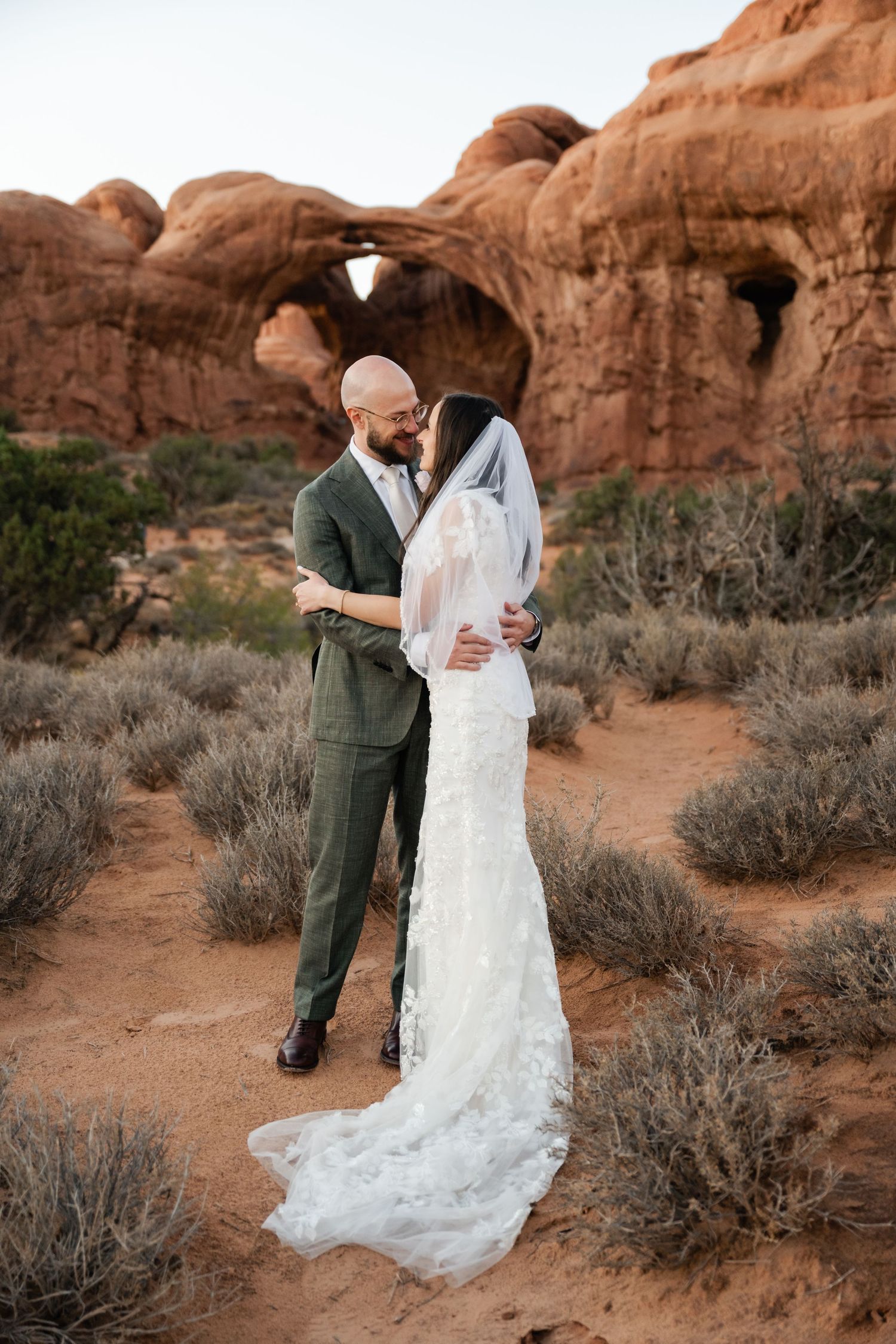 A couple shares an intimate moment among desert brush and red rock formations.