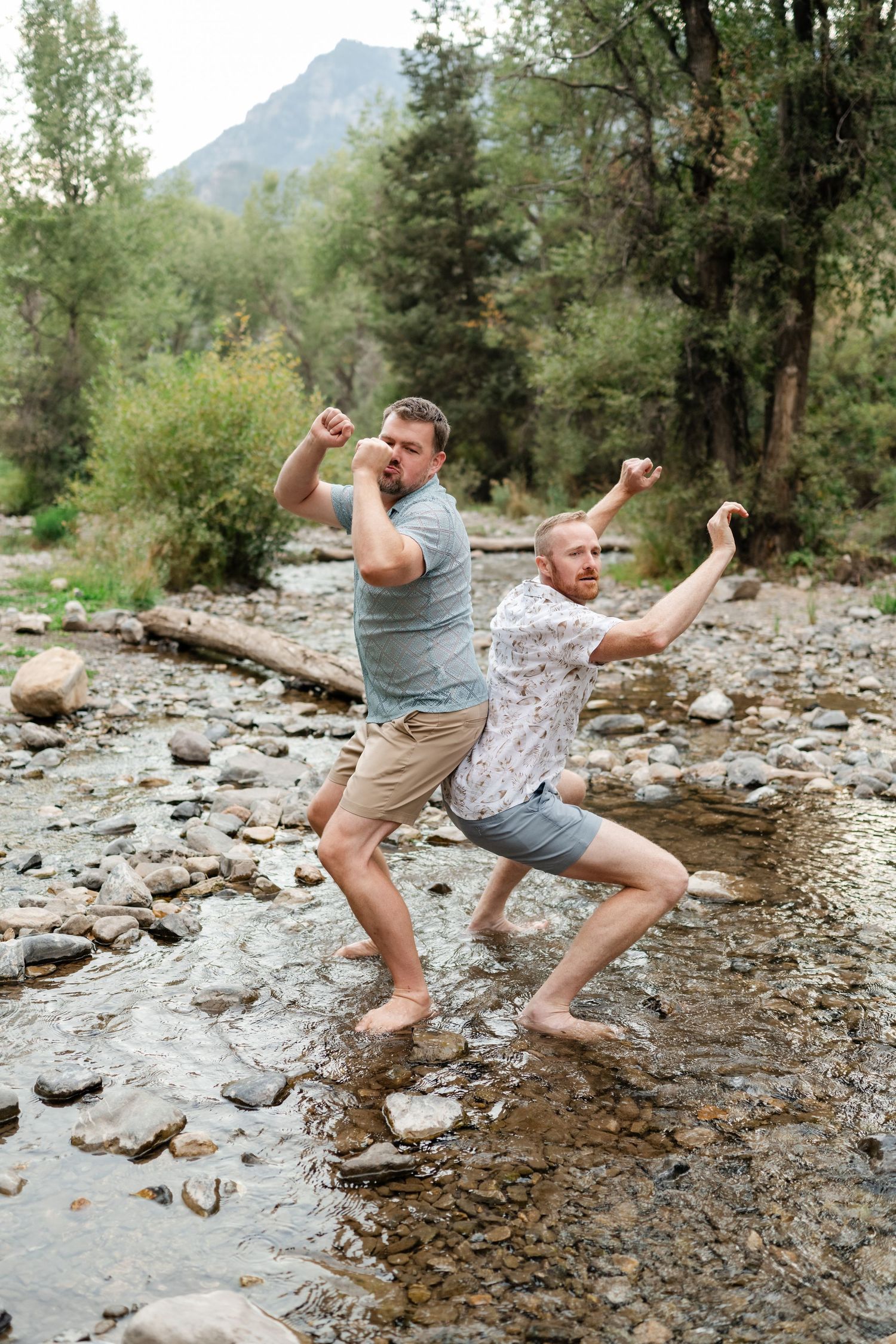 LGBTQ couple engagement photos in the Utah. Two men striking fun poses barefoot in a rocky mountain stream surrounded by lush green trees.
