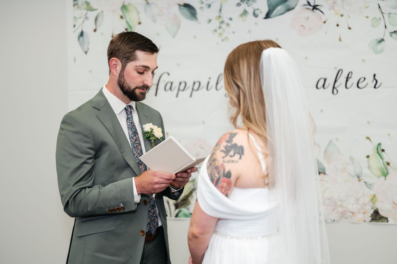 A couple in formal attire looks at their phones during a wedding ceremony against a floral backdrop.