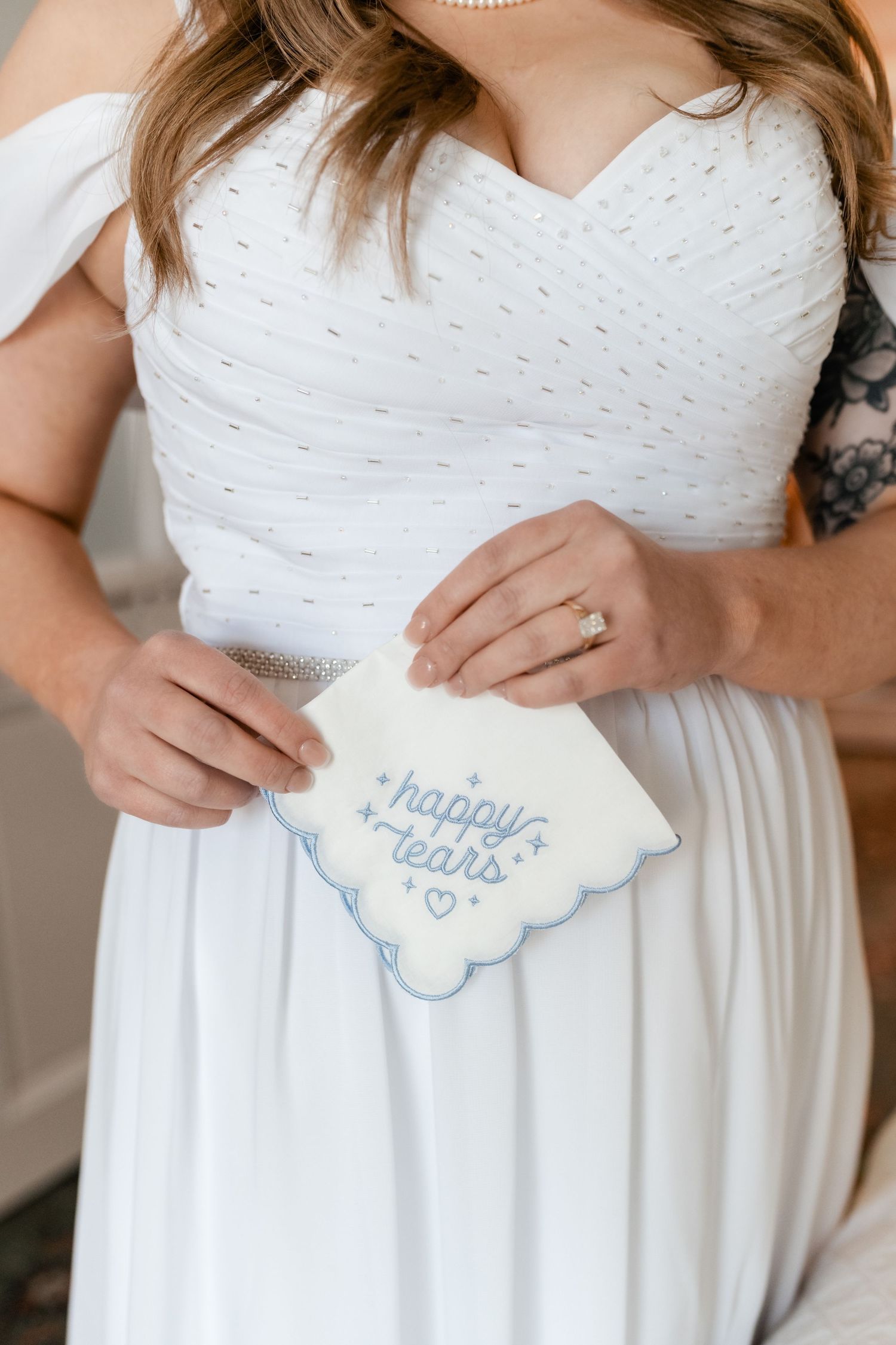 A bride in a white wedding dress holds a decorative handkerchief with 'happy tears' embroidered on it.