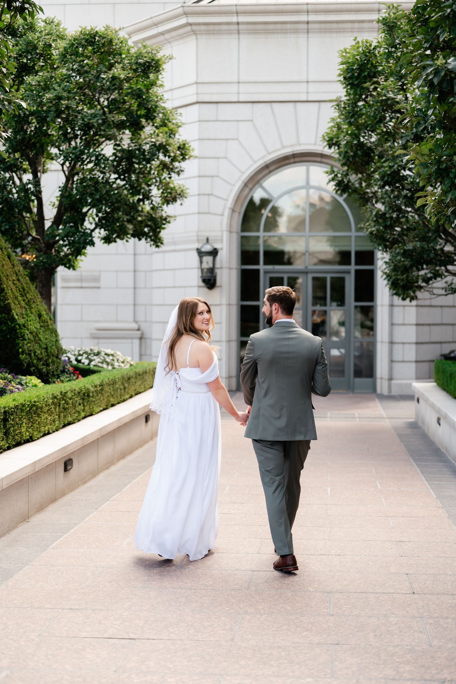 A newlywed couple walks hand in hand along an elegant white stone walkway with arched windows.