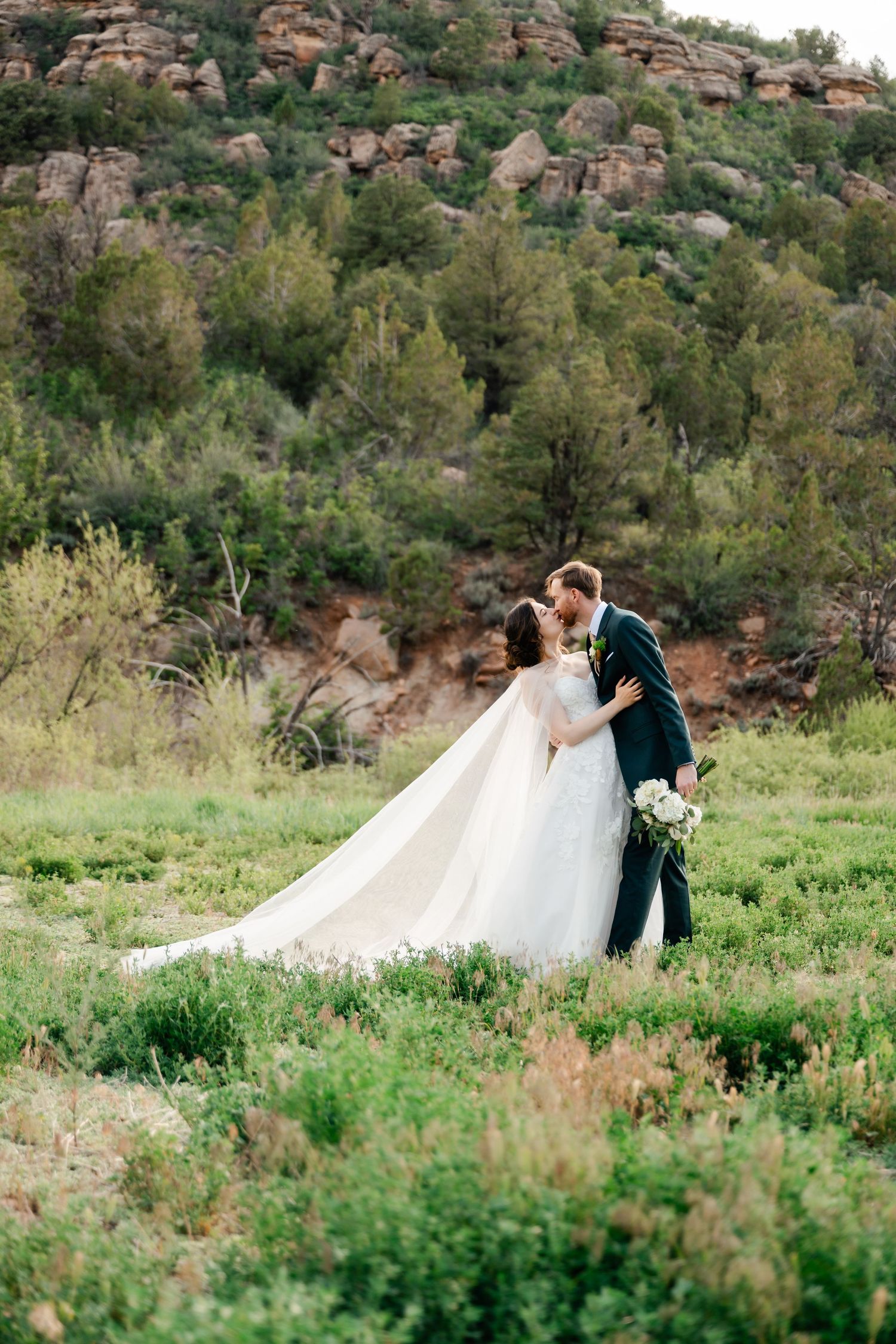 A scenic wedding photoshoot in a green meadow with dramatic red rock formations in the background.