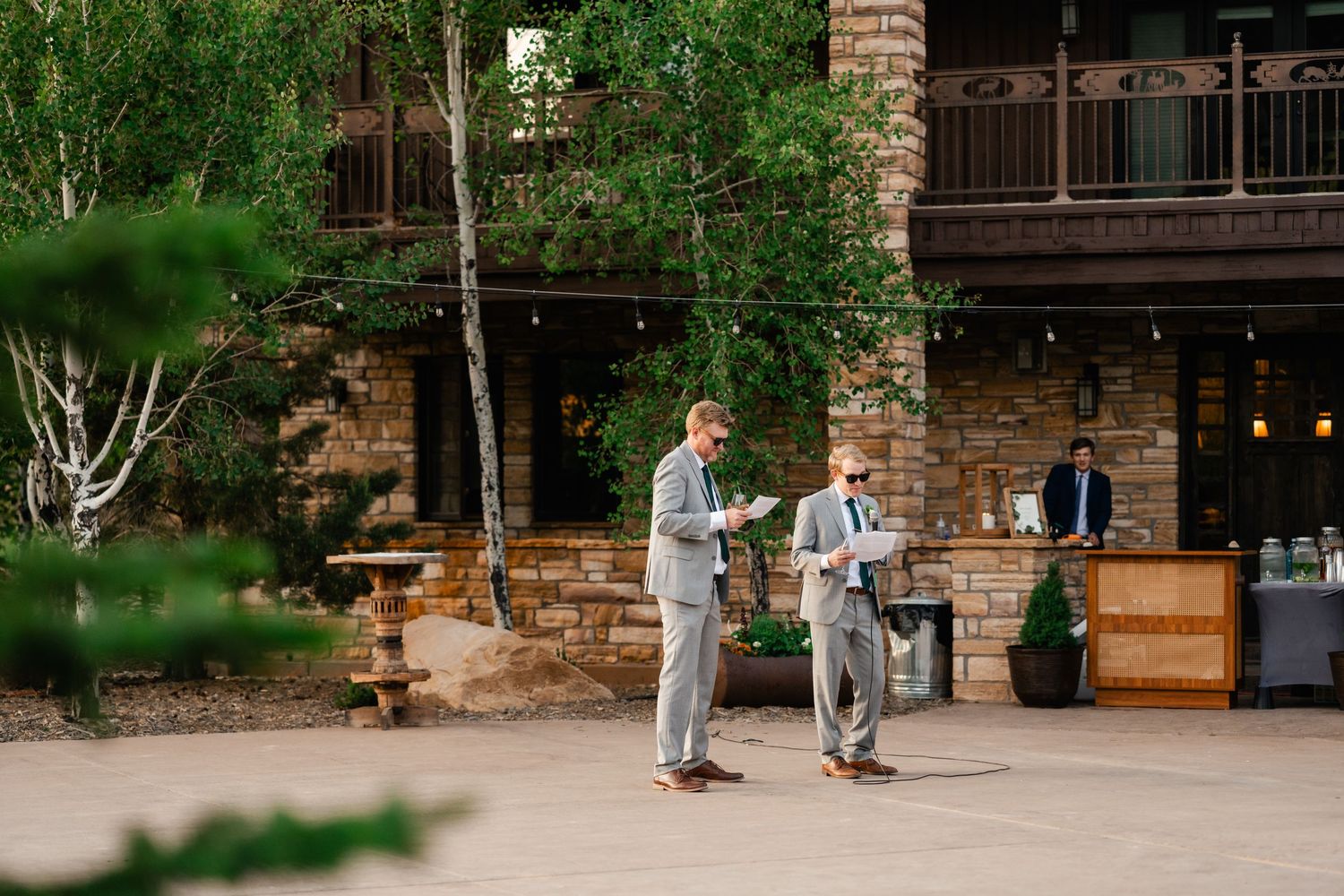 Wedding party walks together on stone pathway outside rustic venue with greenery.