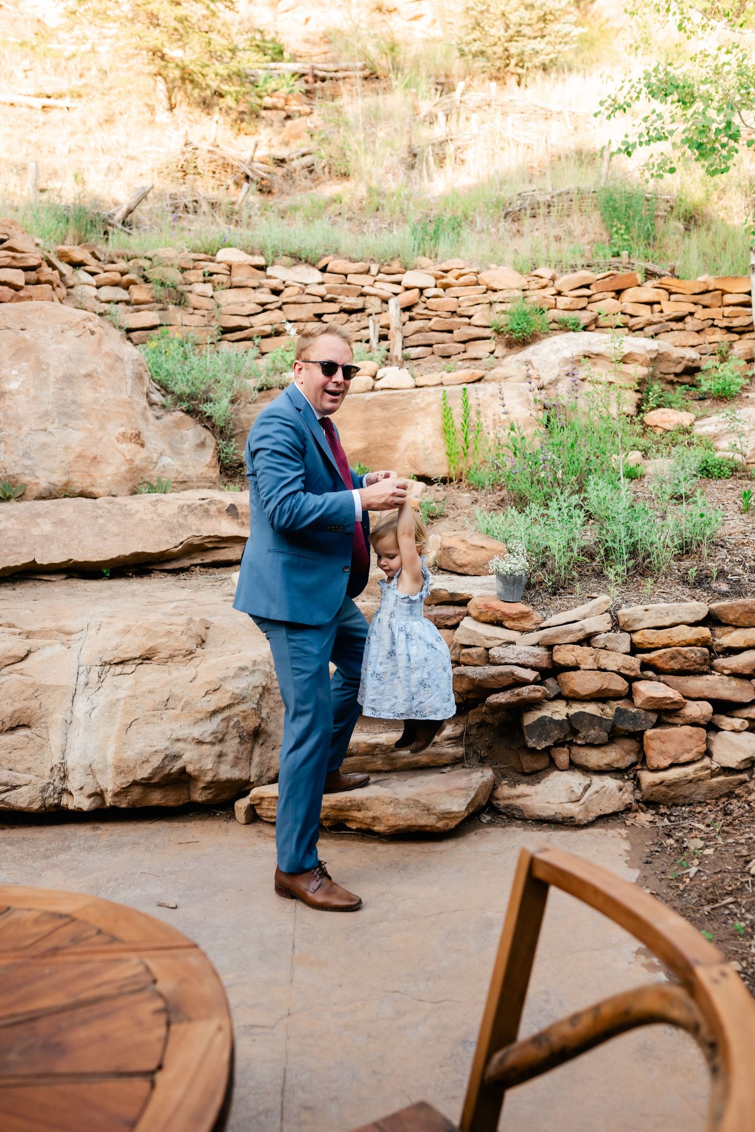 A person in a blue suit dancing outdoors near a rustic stone wall and wooden table.