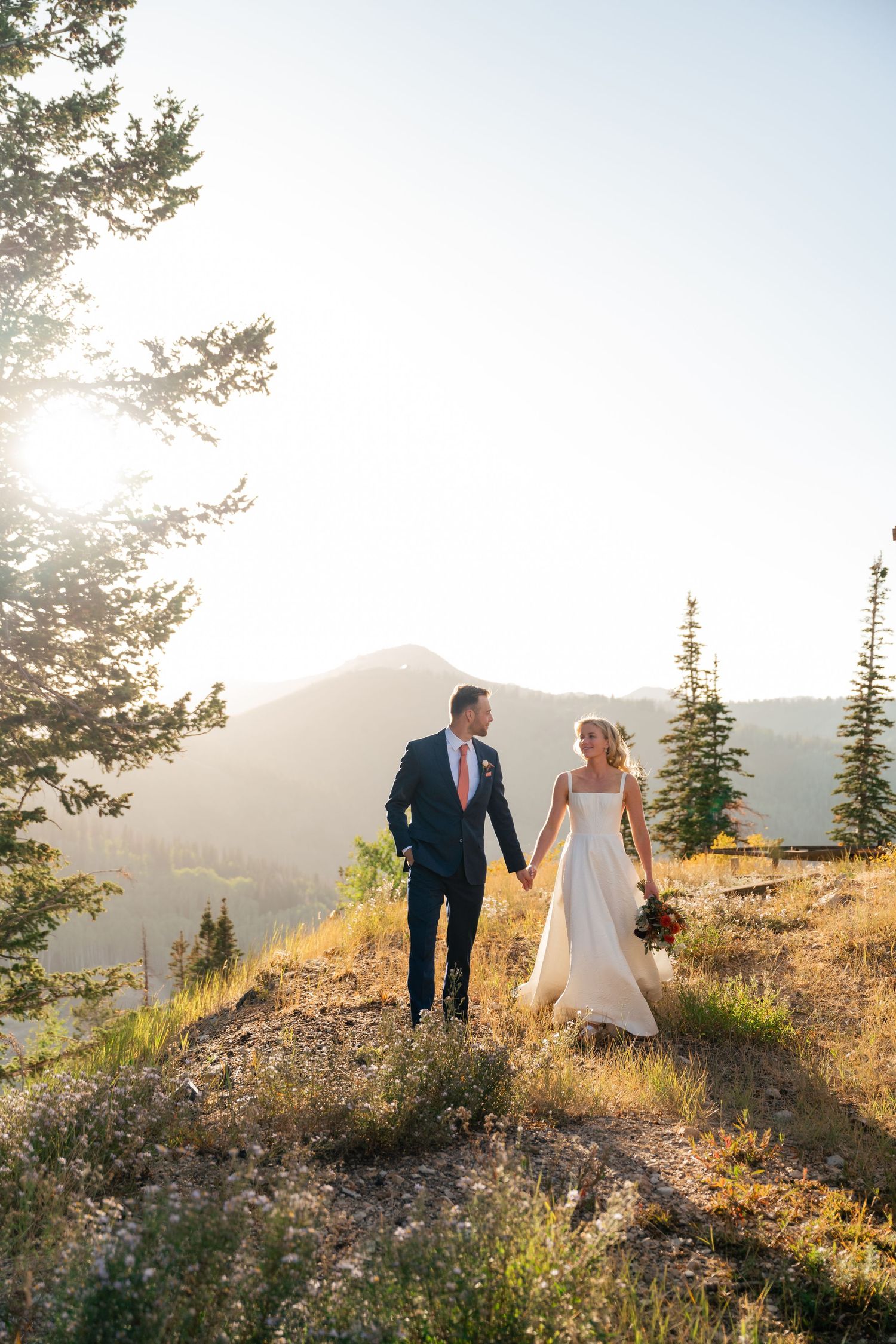 A couple walks hand in hand on a scenic mountain hillside during golden hour with evergreen trees in the background.