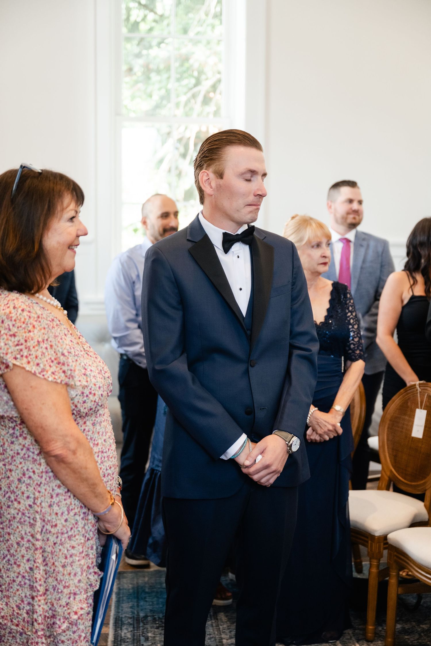 A groom in a black tuxedo stands nervously during a wedding ceremony while guests look on.