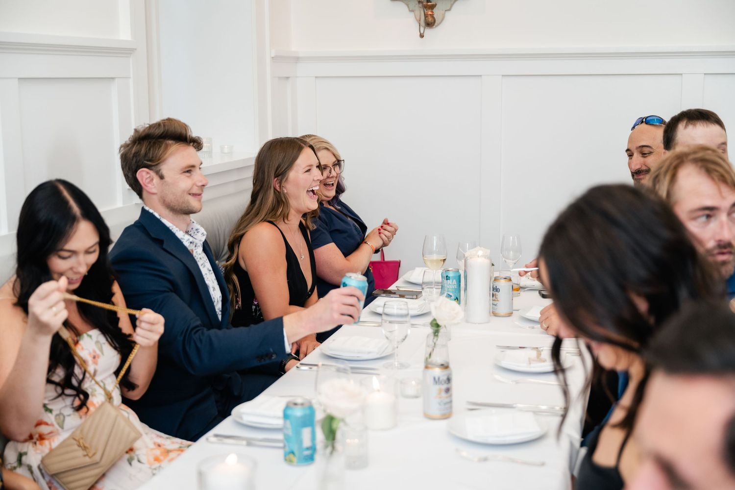 Wedding reception guests enjoying dinner at elegantly set tables in bright white venue.