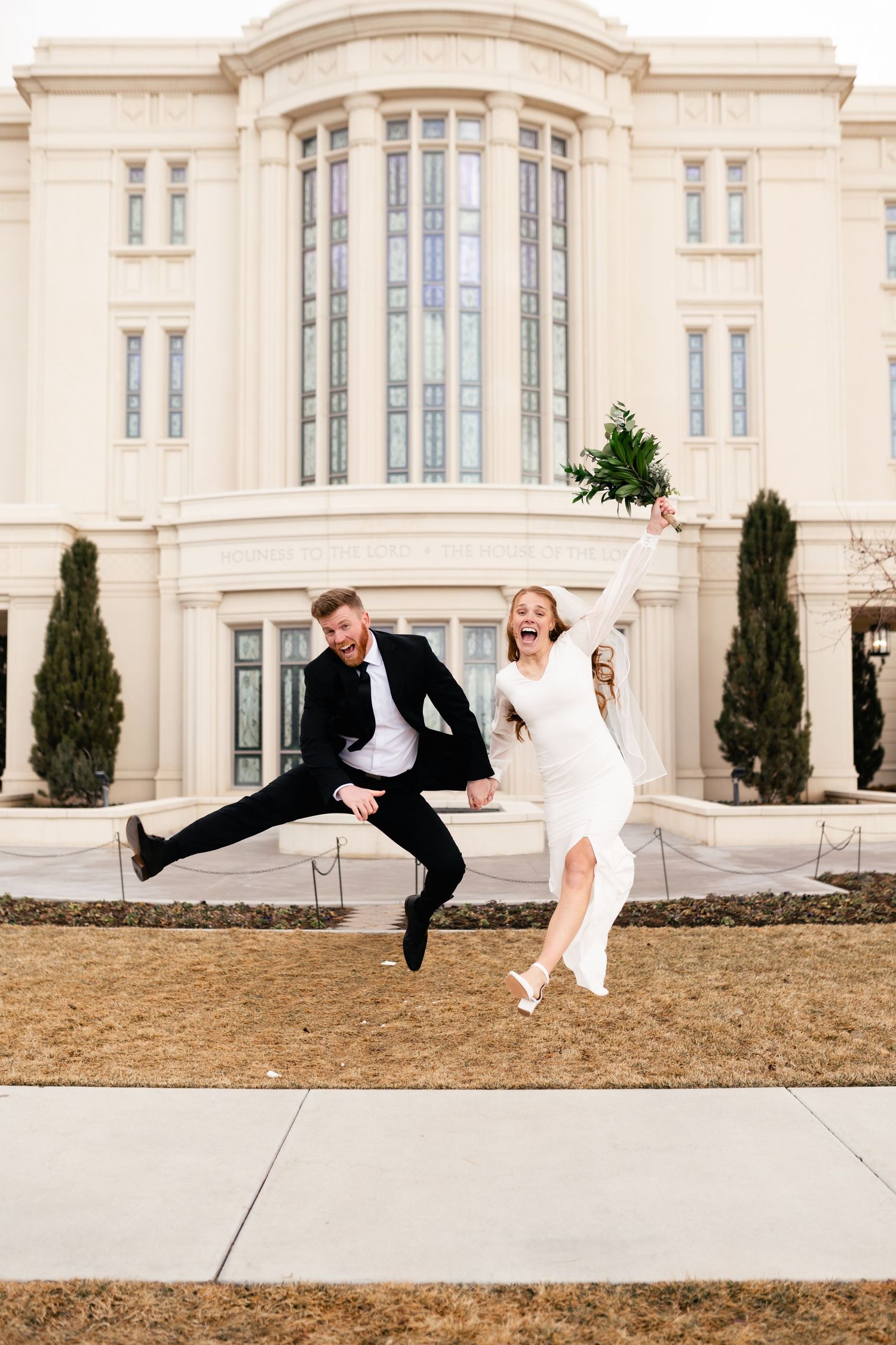 A couple in formal wedding attire playfully leap and dance together in front of a grand historic building. LDS wedding photographer.