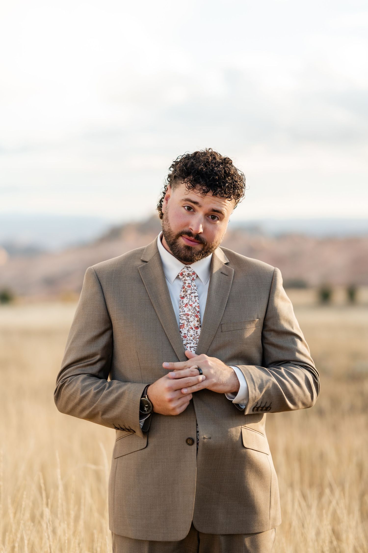 Groom in tan suit and floral tie adjusts his jacket in a golden field.