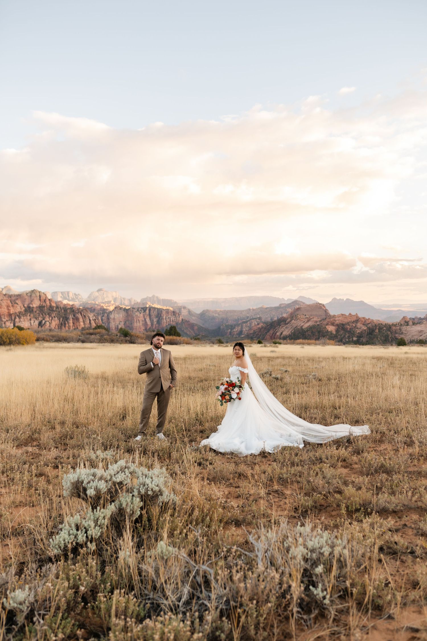 Wedding couple walks through desert brush with dramatic mountain landscape at sunset.