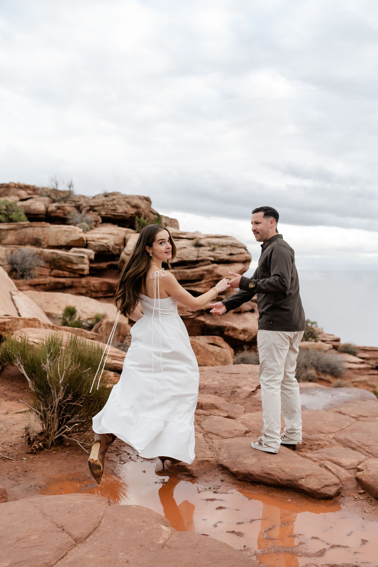 A couple dances playfully on red sandstone rocks against a cloudy sky backdrop.