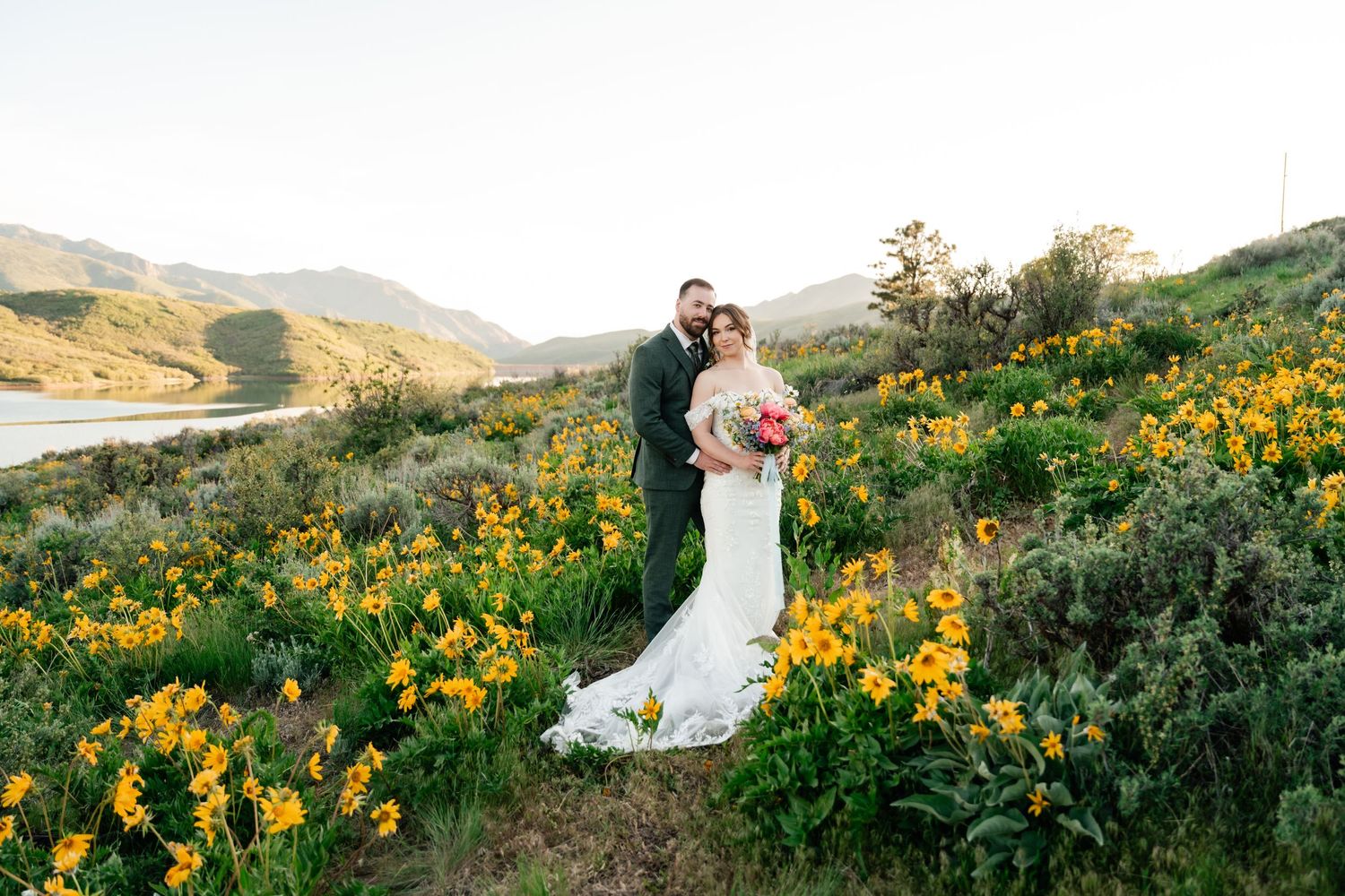 Newlywed couple embraces in scenic mountain meadow filled with vibrant yellow wildflowers at sunset.