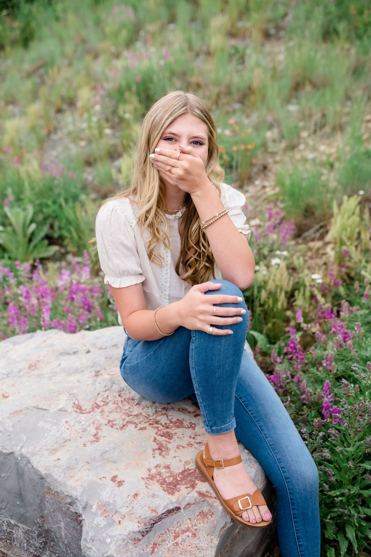 Someone in a white blouse and jeans sits on a rock surrounded by pink wildflowers in a natural outdoor setting.