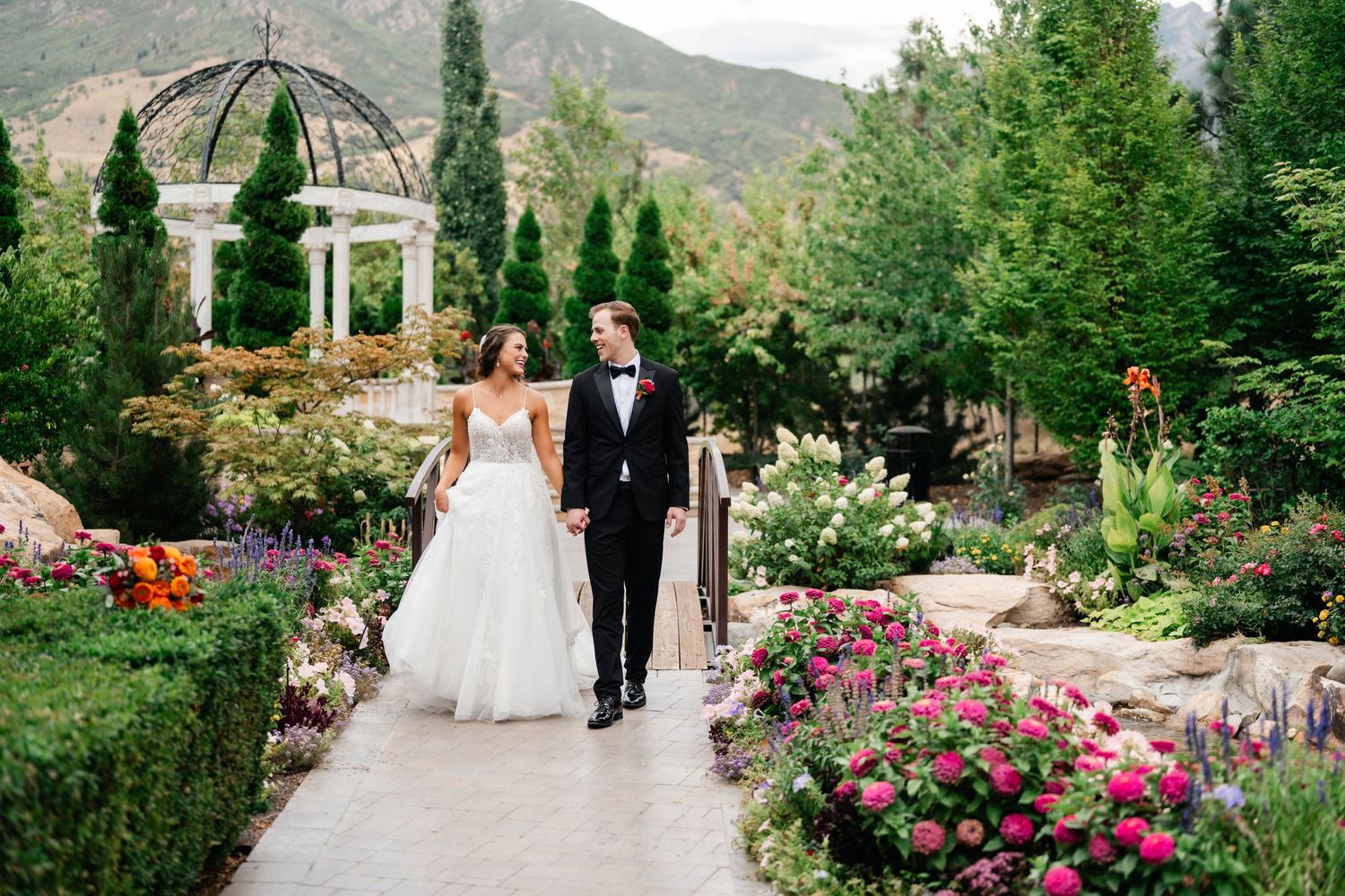 A couple walks through a lush garden with a stone gazebo and colorful flowers at Bellissimo Gardens. Best Utah Wedding Photographers.
