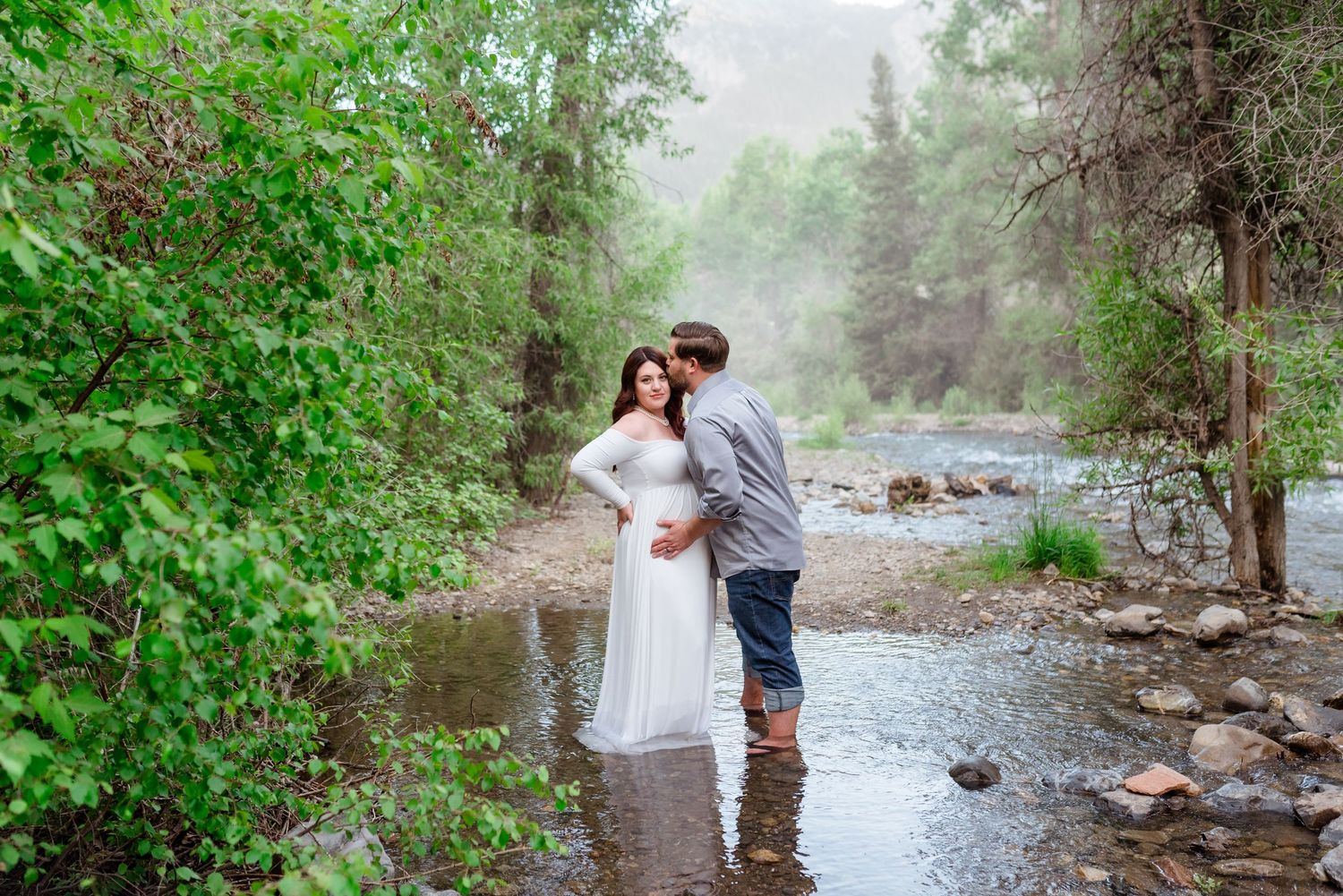 Two people embrace by a misty river in a lush green forest setting during a romantic outdoor photoshoot.
