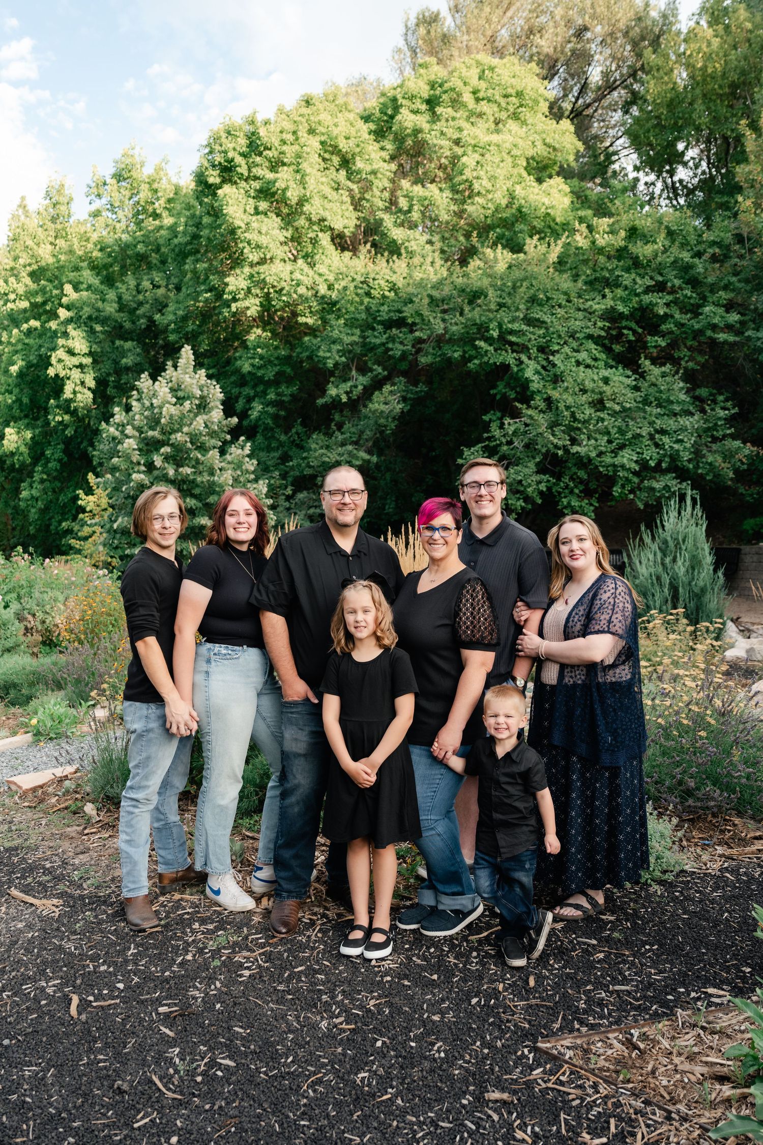 A group of people wearing black and casual clothing pose together outdoors in a garden setting.