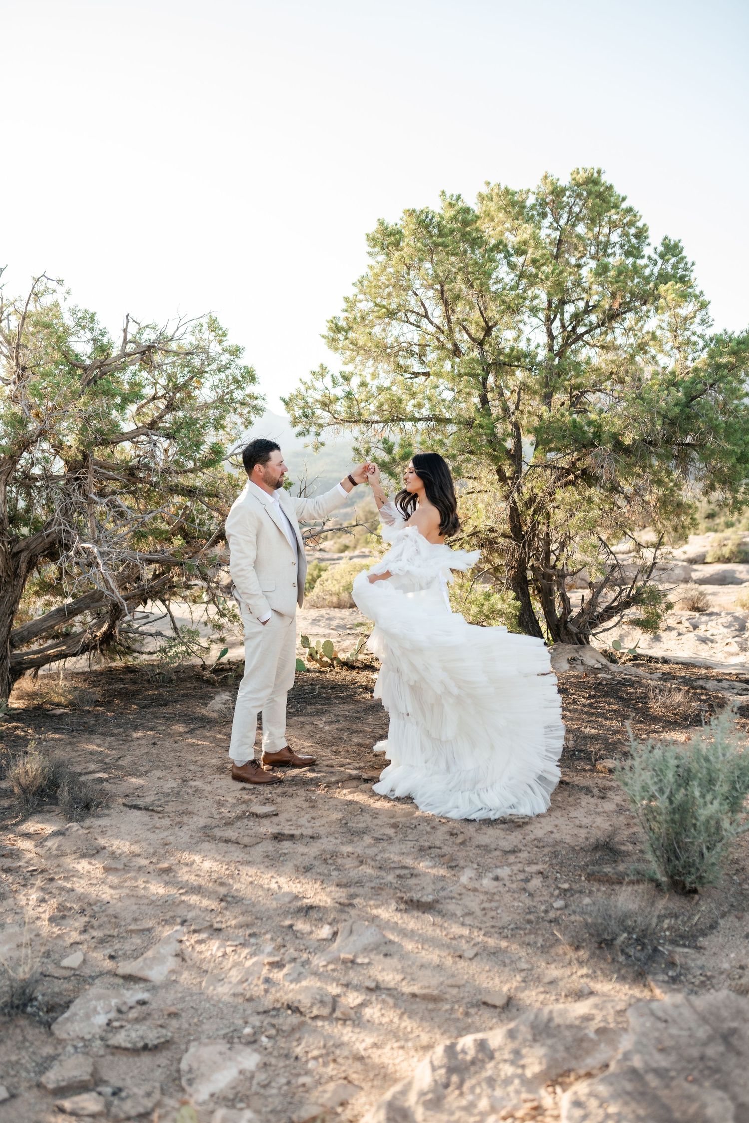 Zion destination photographer, Dancing couple in white attire twirls among desert brush and juniper trees.