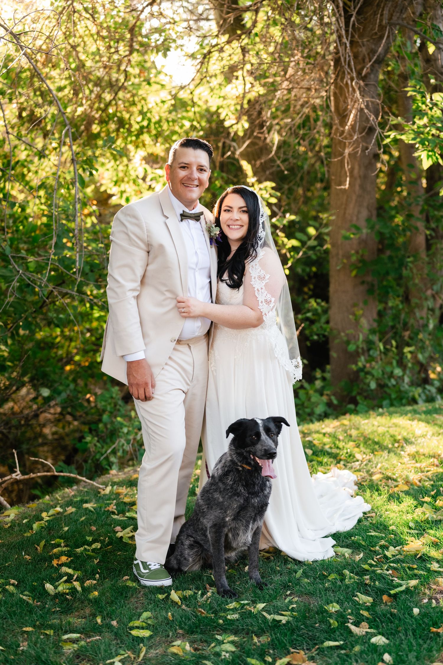 A couple in white wedding attire poses with their black dog in a sunlit garden setting.