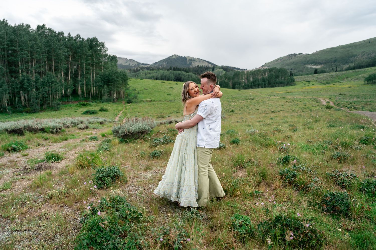 A couple embraces in a sage green meadow surrounded by mountains and aspen trees in Colorado. Utah mountain engagement session