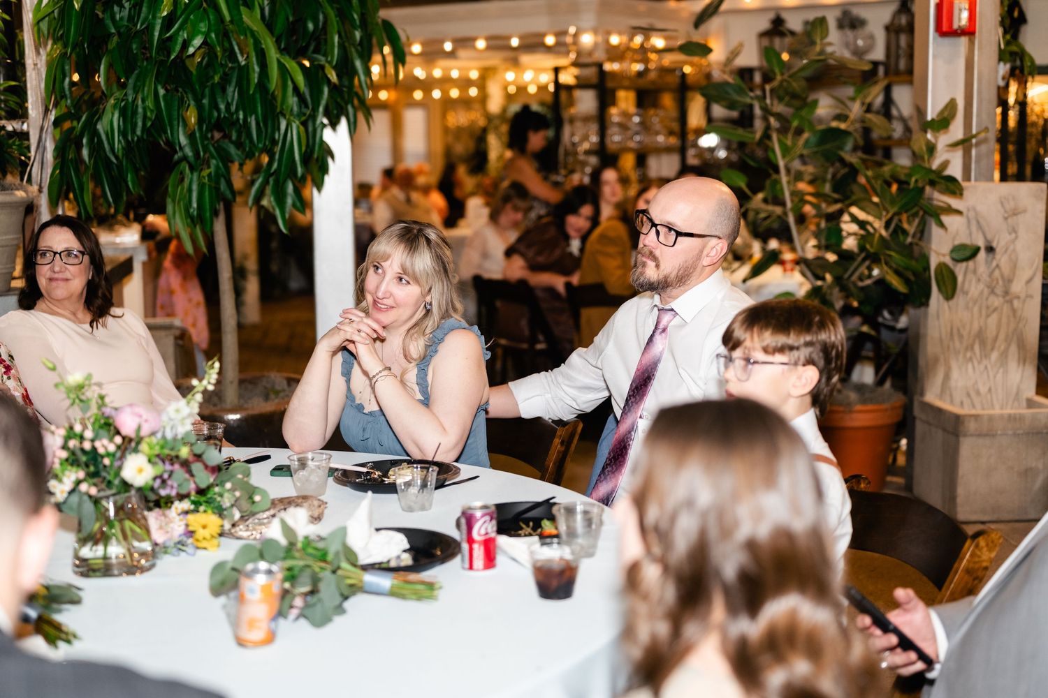 Wedding reception guests seated at decorated table with string lights and greenery in a restaurant setting.