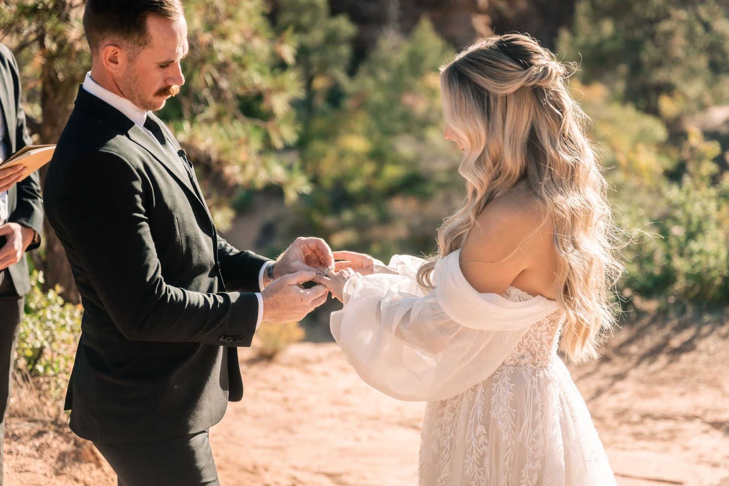A couple exchanges rings during an intimate outdoor wedding ceremony against a sunlit natural backdrop.