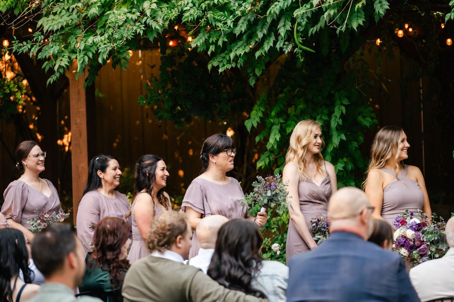 Wedding party members in pink dresses stand together during an outdoor garden ceremony.