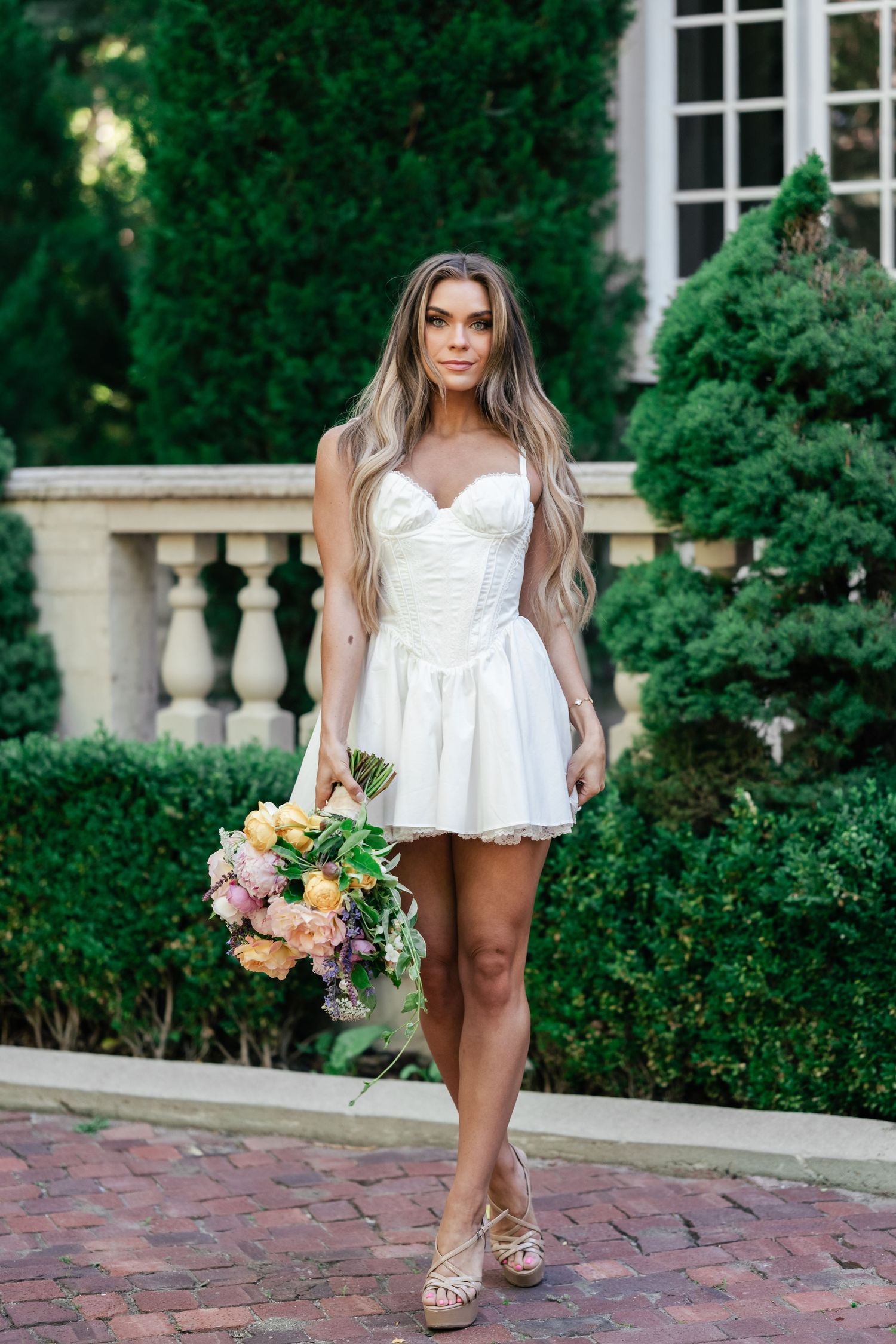 Someone in a white strapless mini dress holds a pastel bouquet while standing on brick pavers near stone balustrades.