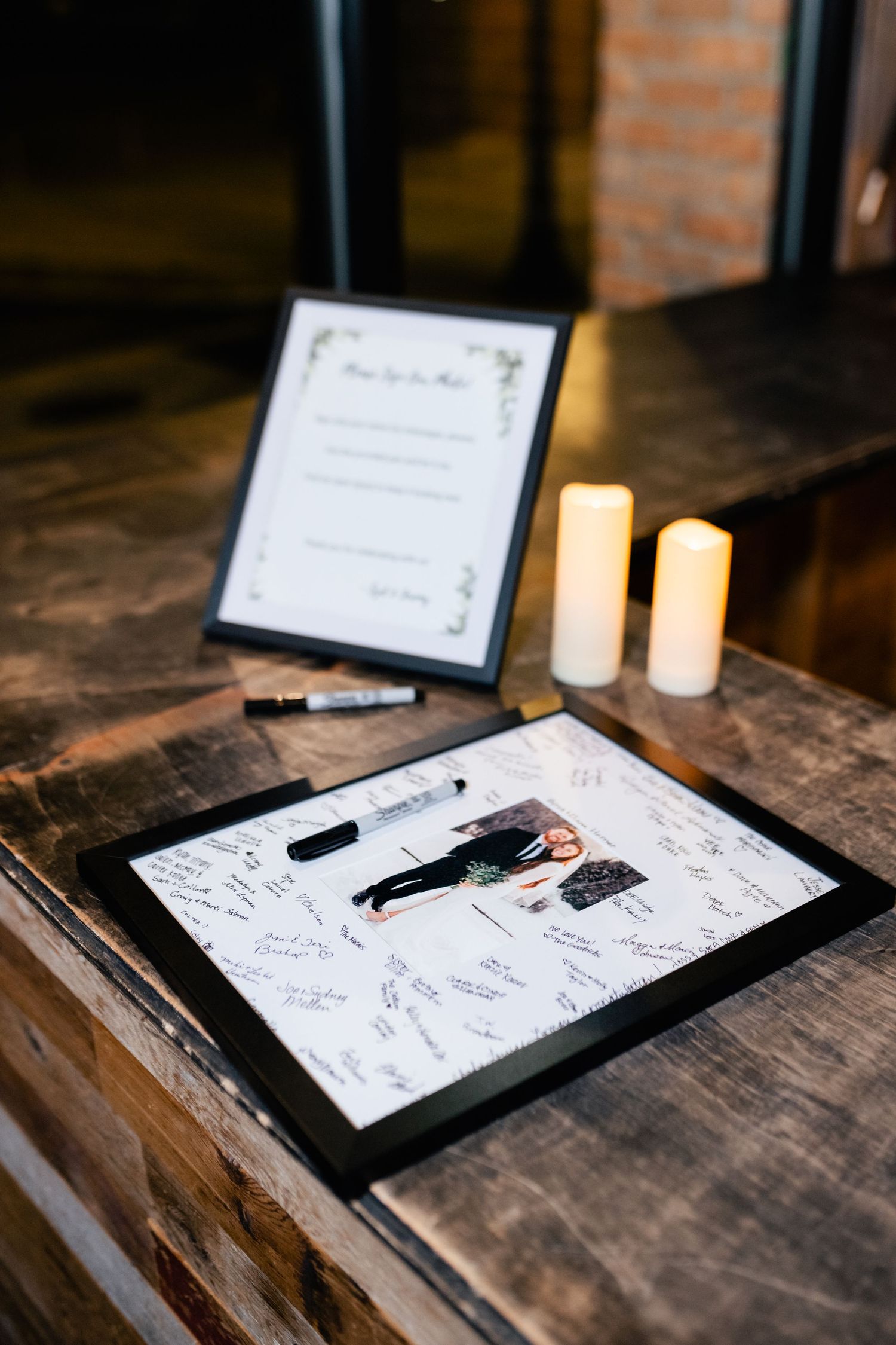 A wedding guest book with signatures lies on a rustic wooden table next to glowing candles and pens.
