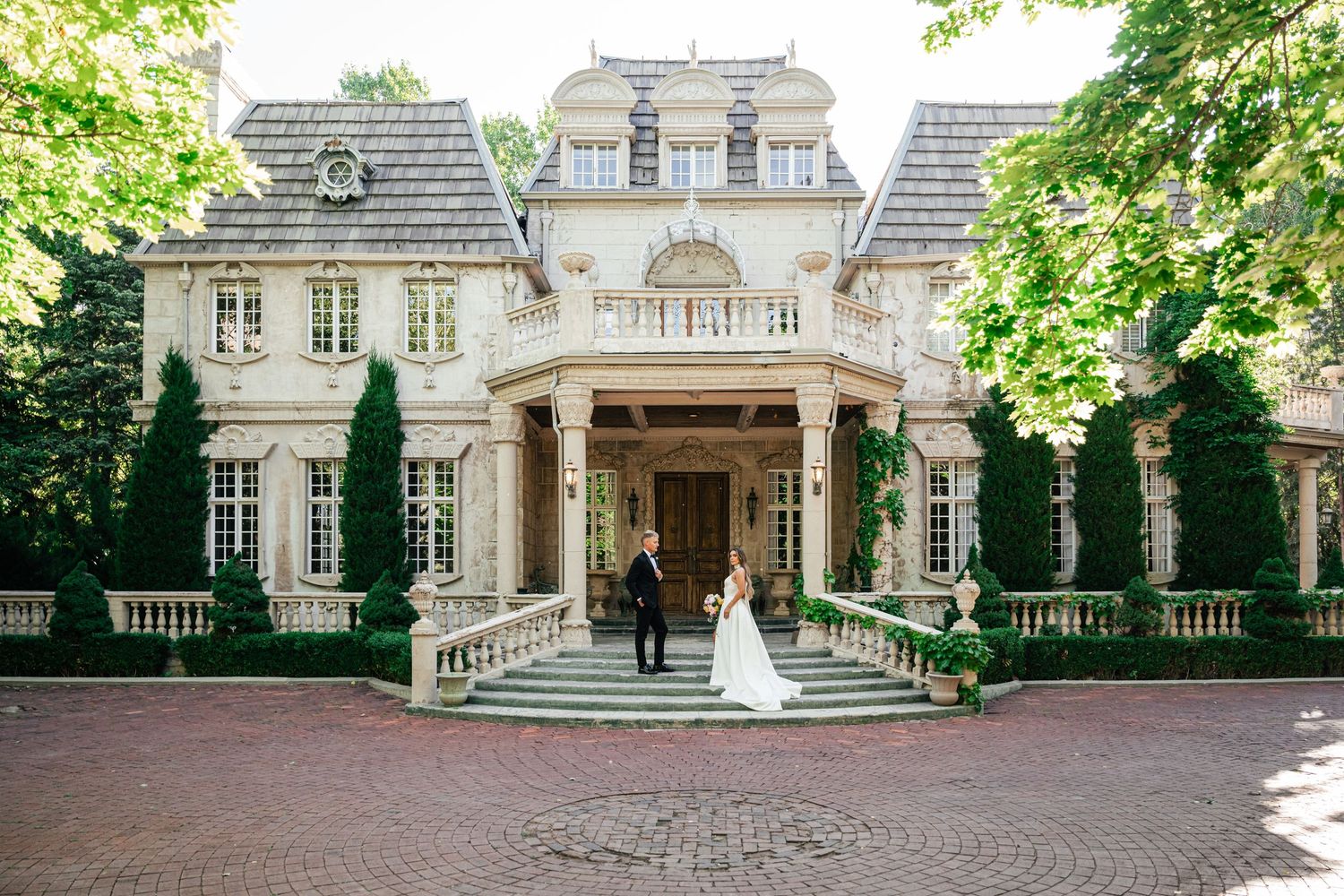 Elegant wedding panorama showing a classic mansion with grand architecture and stately columns framed by lush green trees.