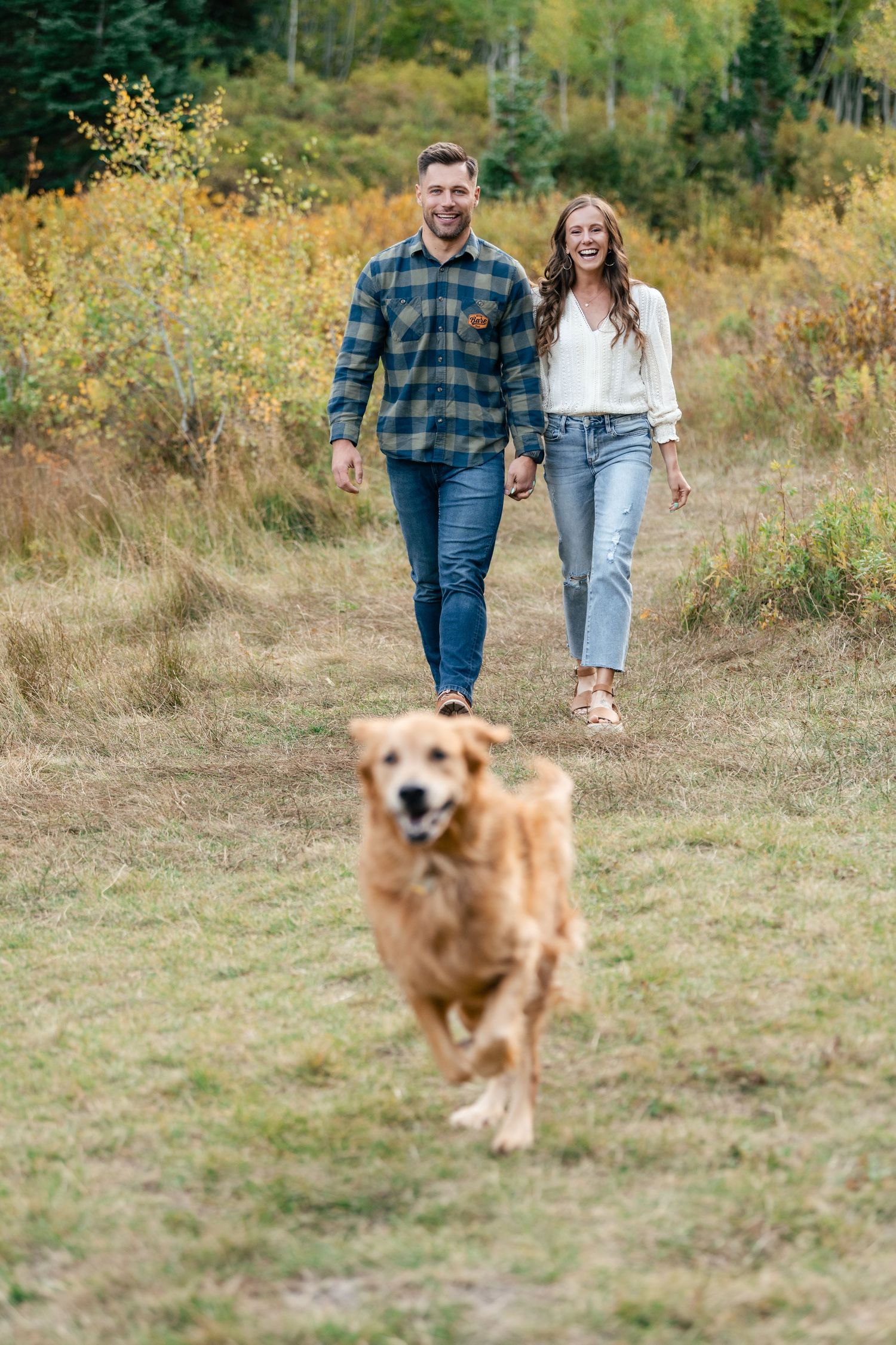 A happy Golden Retriever running on a trail ahead of two people walking together on an autumn day.