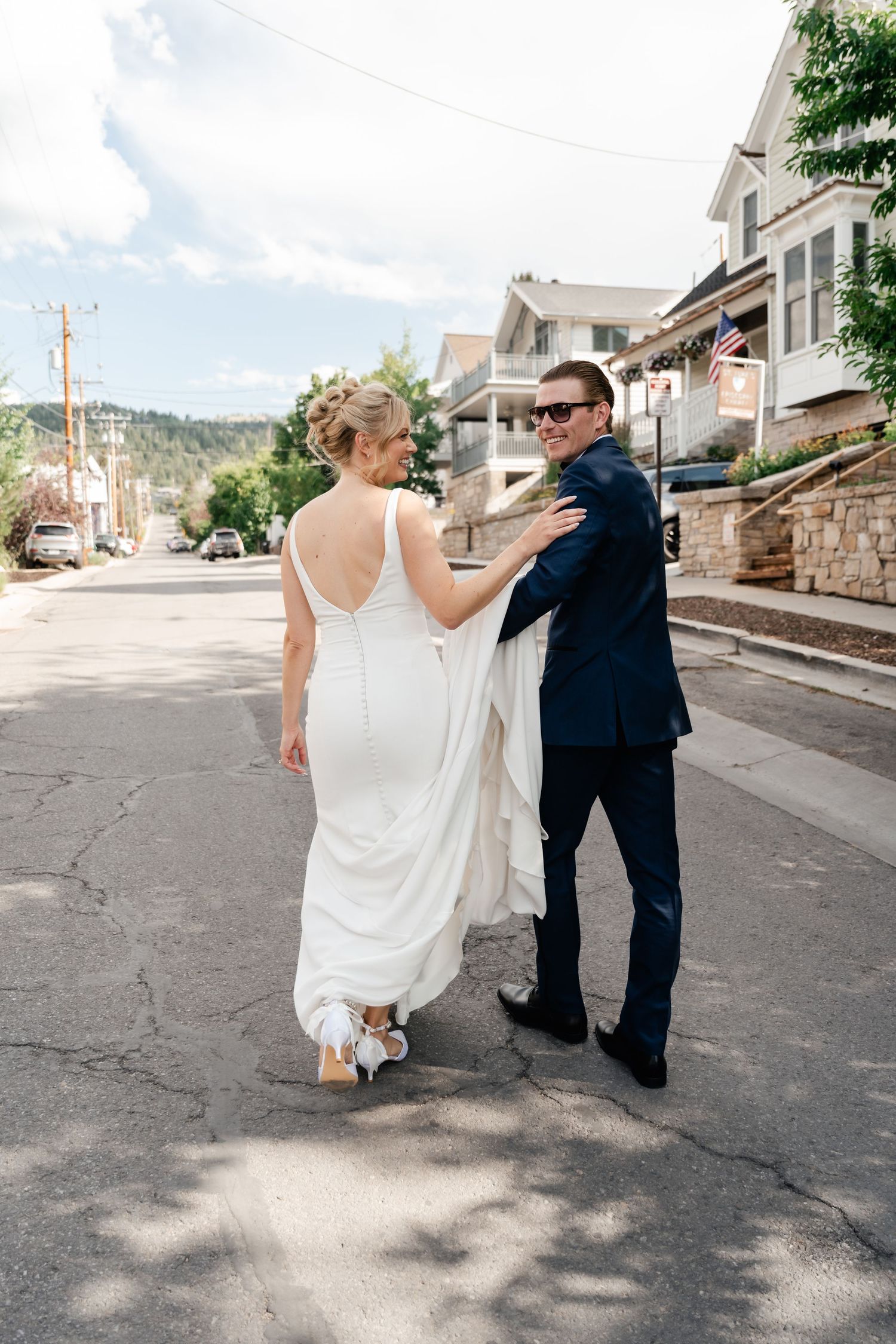 Bride in white gown and groom in navy suit walk down a sunny street, smiling at each other on their wedding day in Park City.