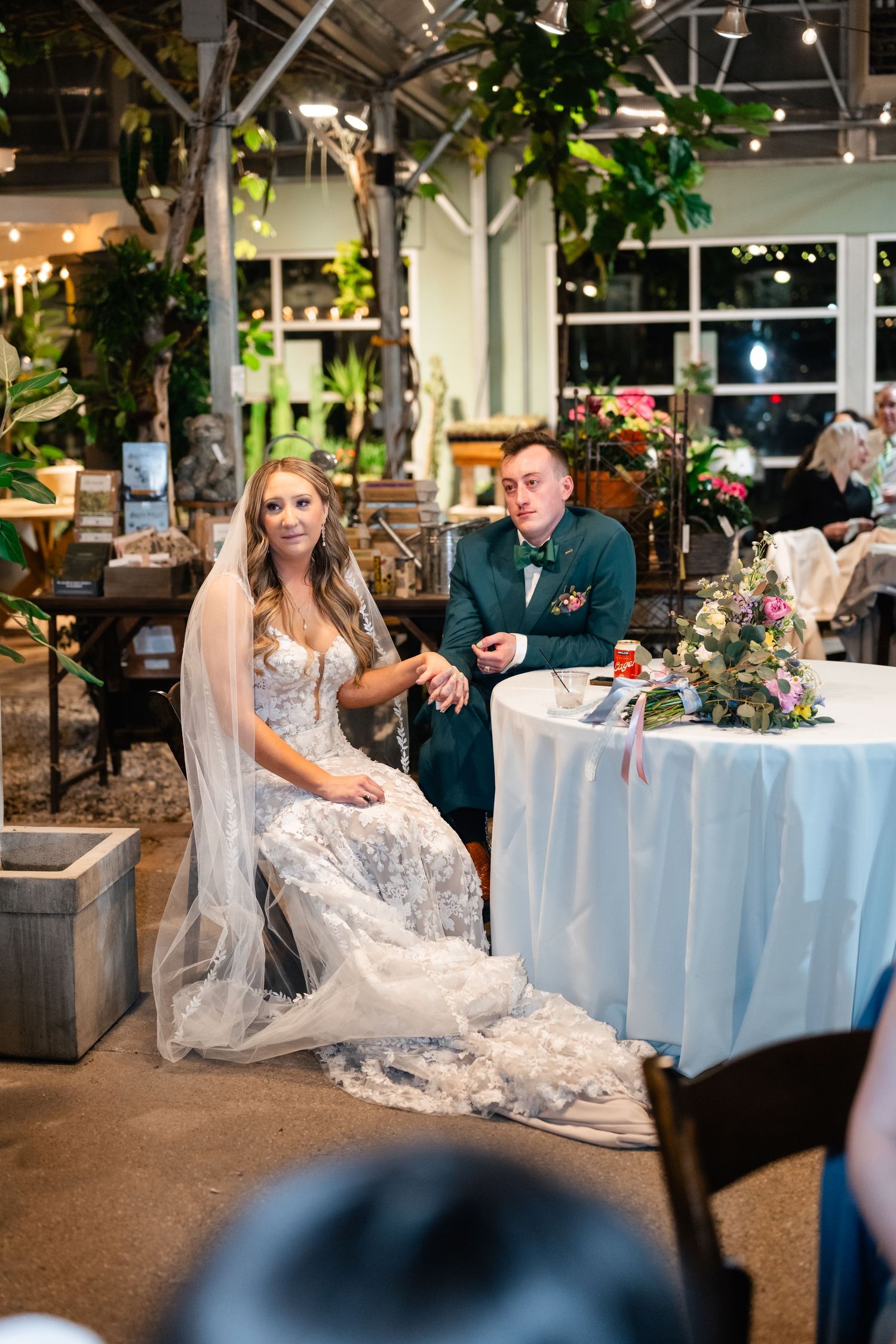 A couple sits at a decorated table during a wedding reception in a modern venue with indoor plants.