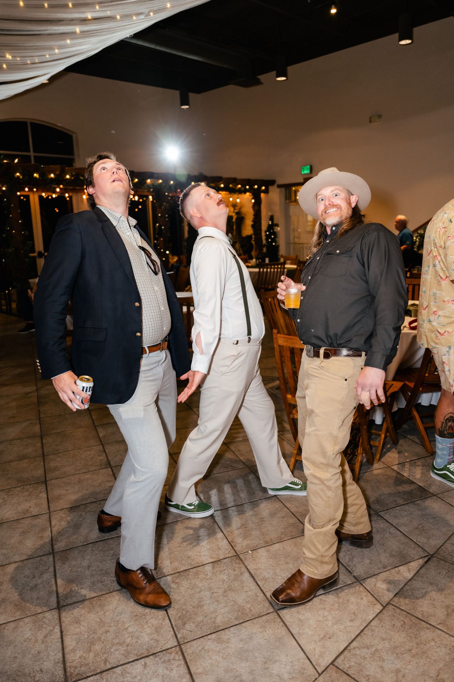Three guests at an indoor party strike animated poses on a tiled dance floor under string lights and ambient venue lighting.