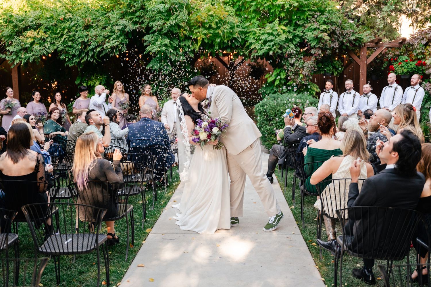 A couple kisses at their outdoor garden wedding ceremony while guests blow bubbles. Alpine Art Center. Utah Wedding Photographer.