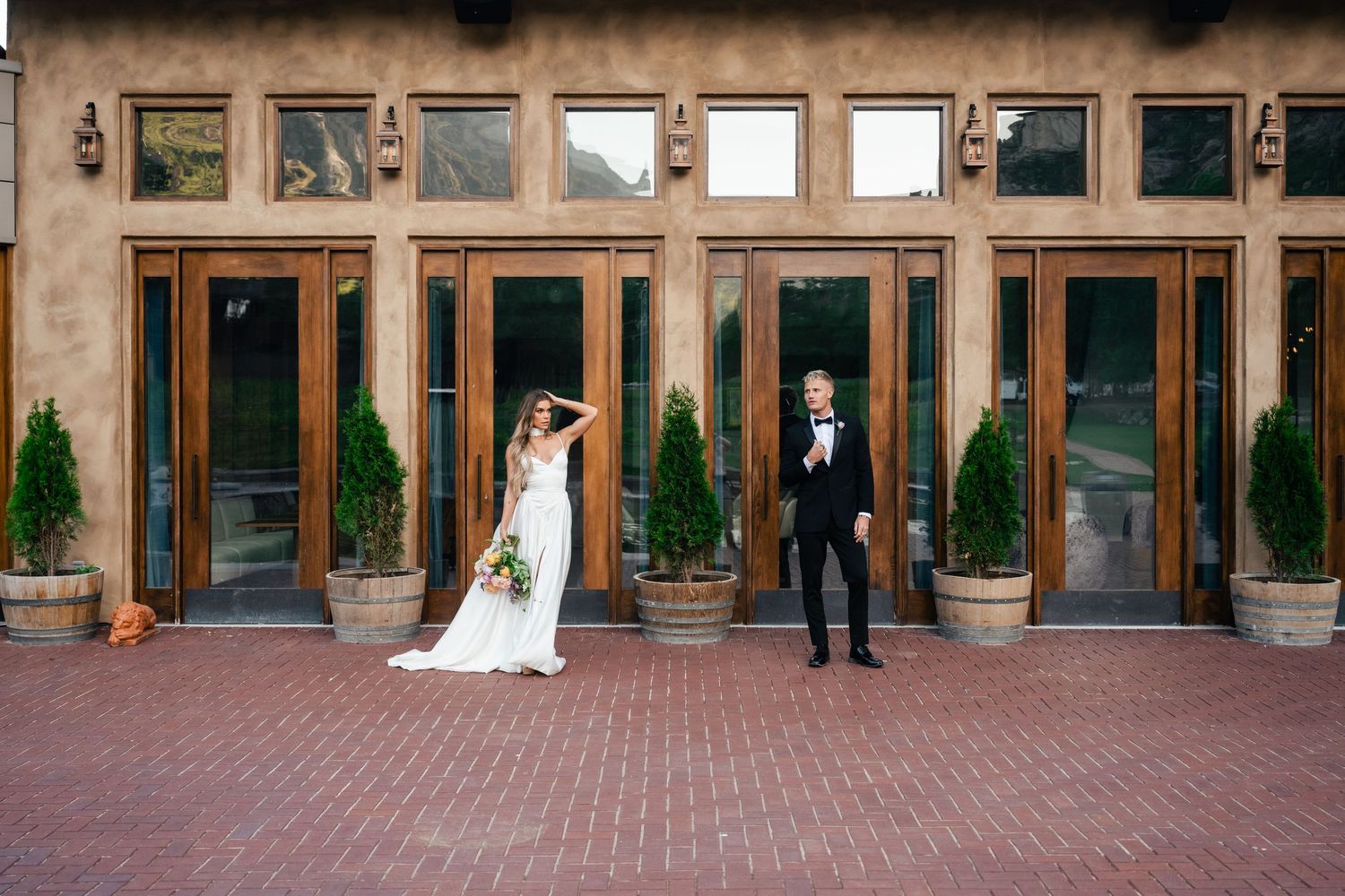 Elegant wedding couple posed separately against rustic glass and wood exterior.