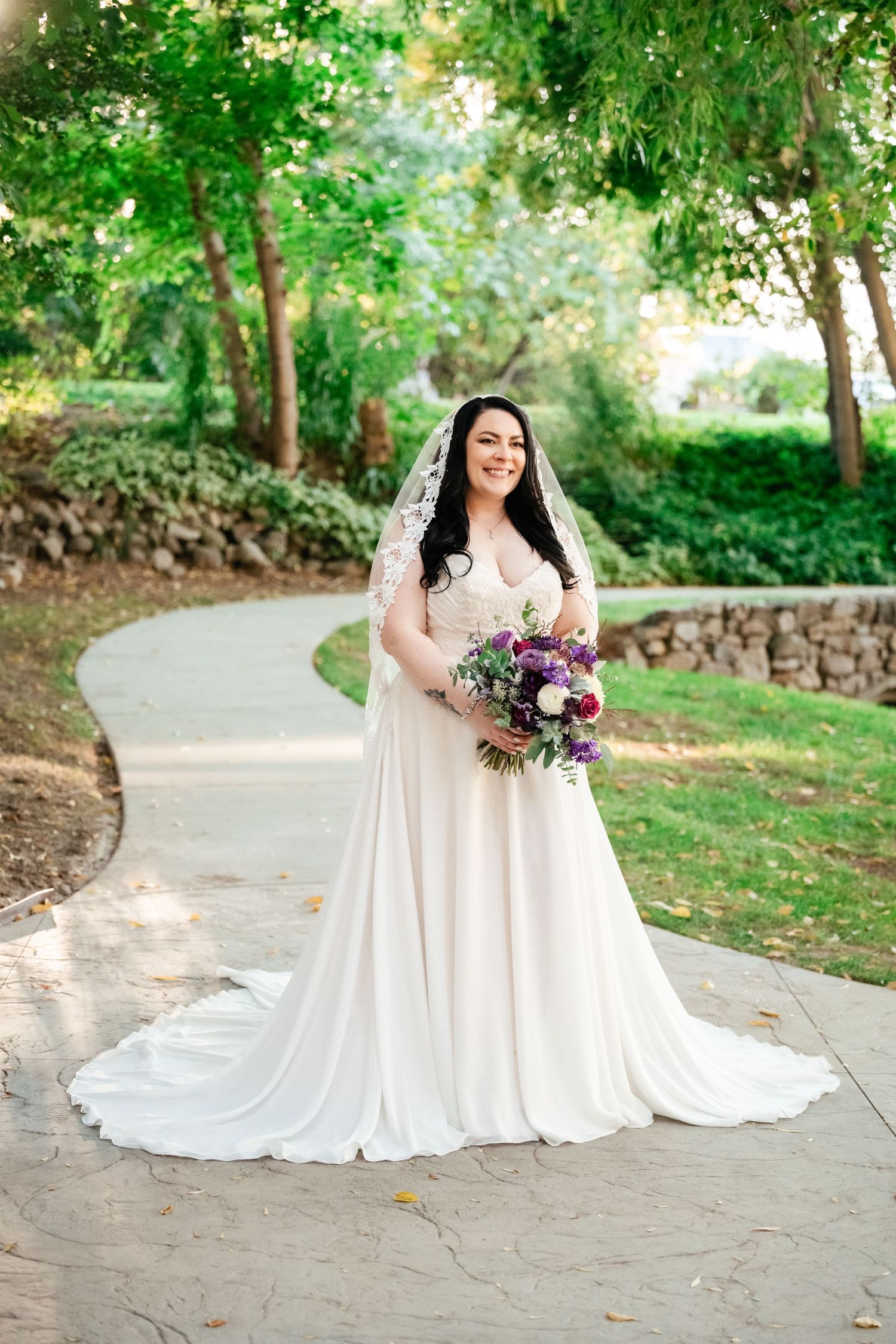 A bride in a flowing white dress and veil poses with a vibrant purple and burgundy bouquet on a garden path.