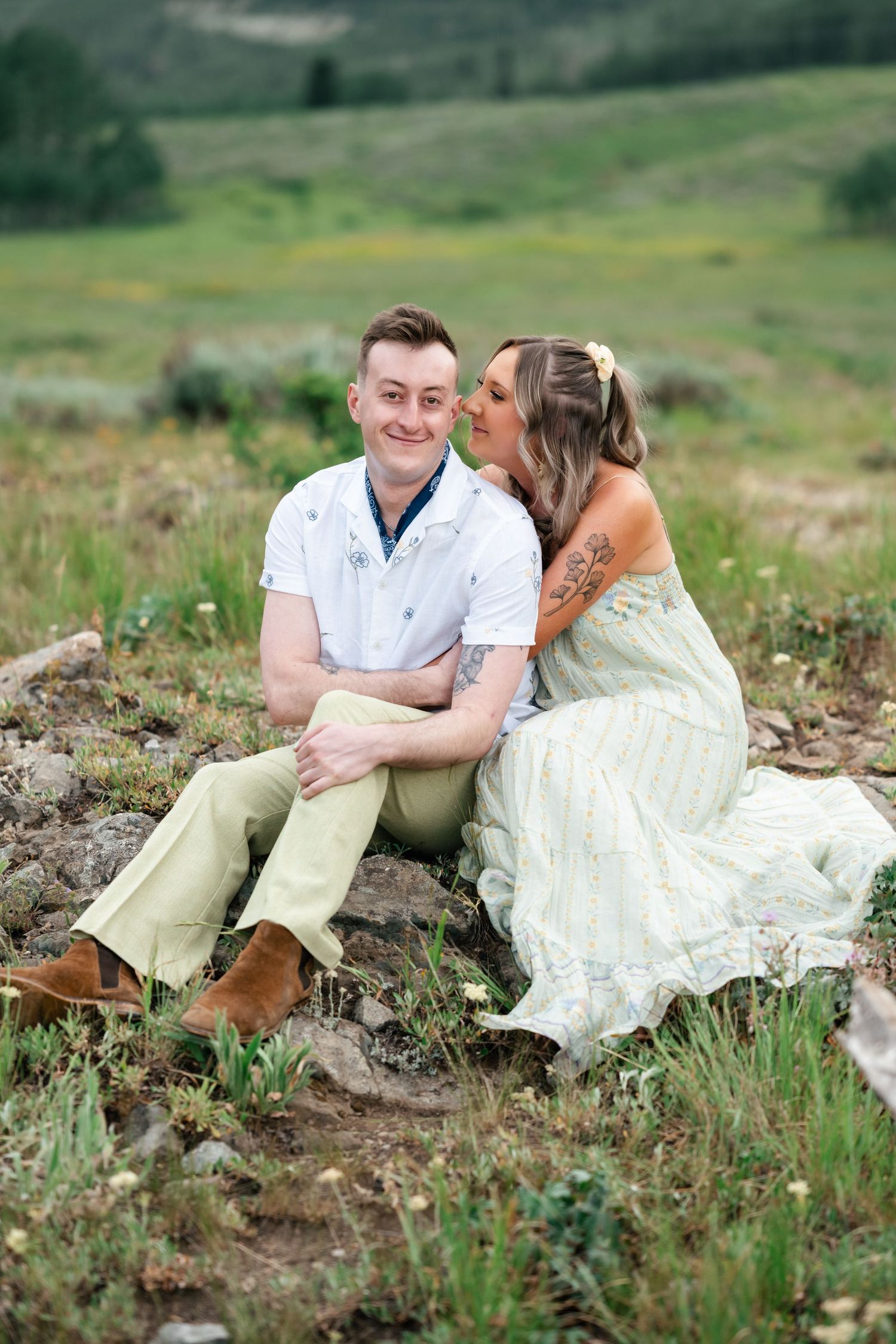 A couple in casual and wedding attire sharing a tender moment while sitting in a green mountain meadow. Utah mountain engagement session