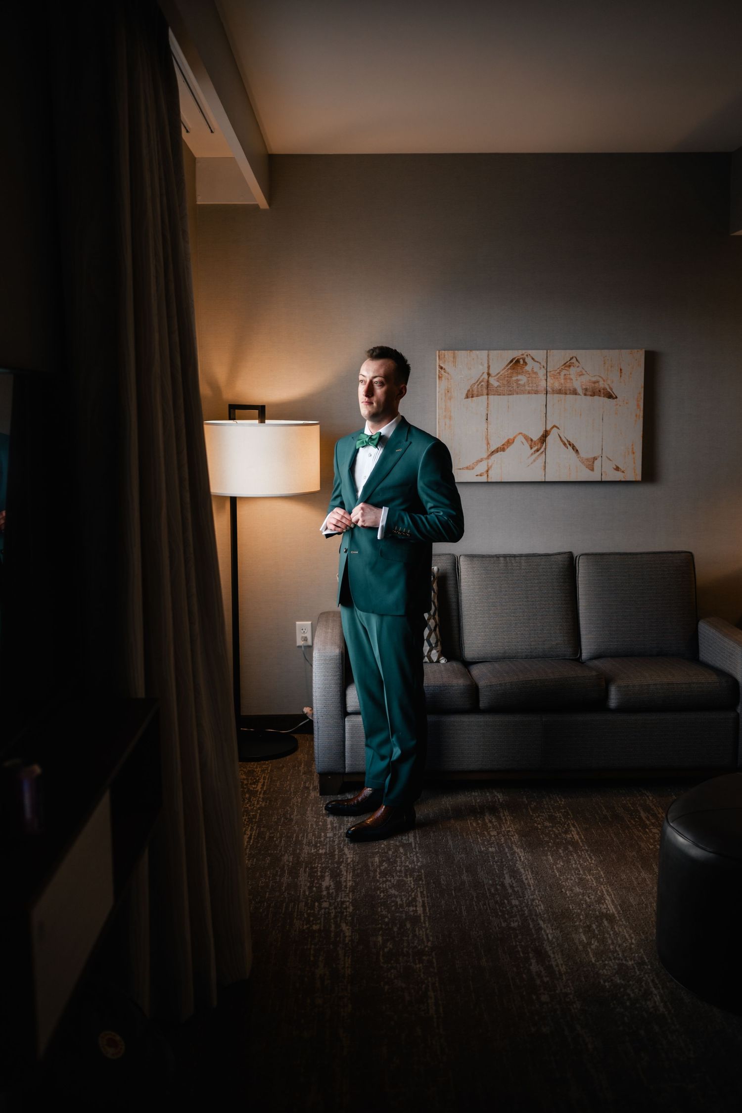 A person wearing a dark teal suit stands in a dimly lit hotel room with modern decor and warm lighting.