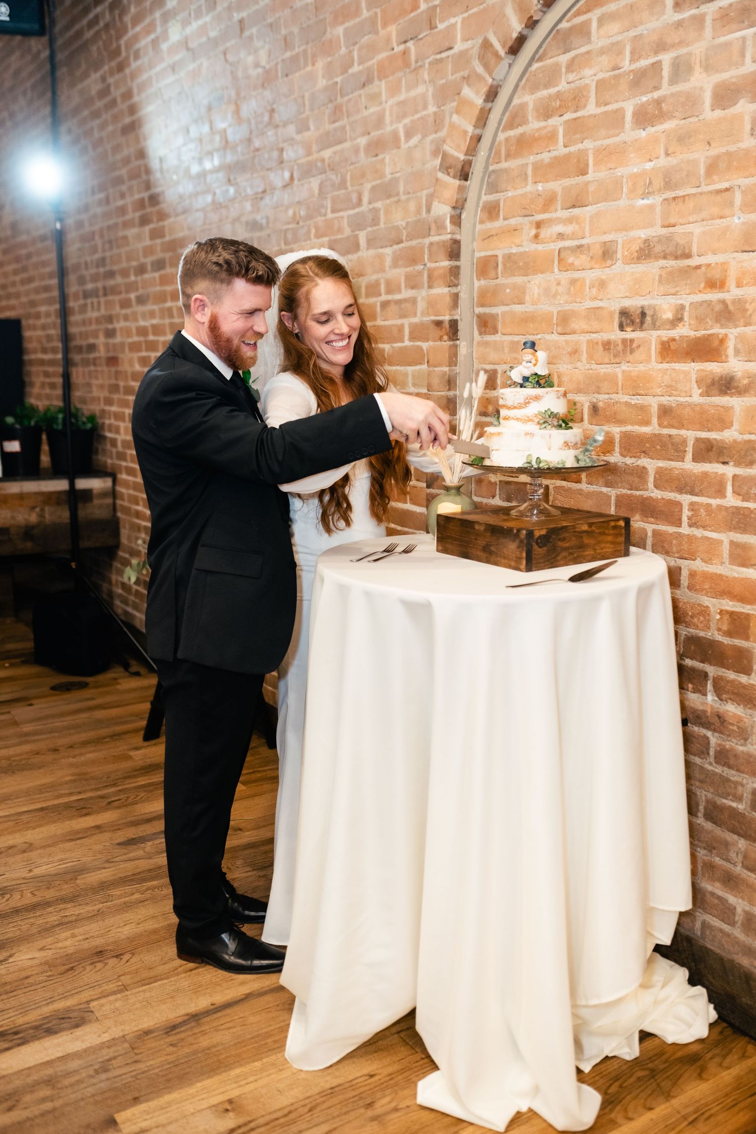 A couple in wedding attire cuts their white tiered cake together at a rustic brick venue.