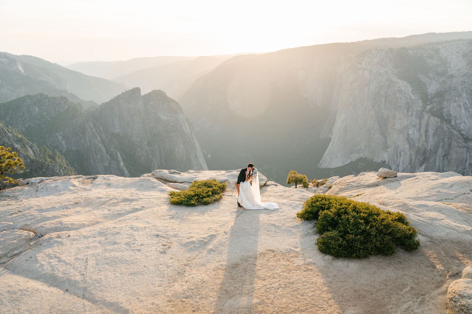 Bride and groom embrace on granite overlook at sunset with sweeping Yosemite valley views.