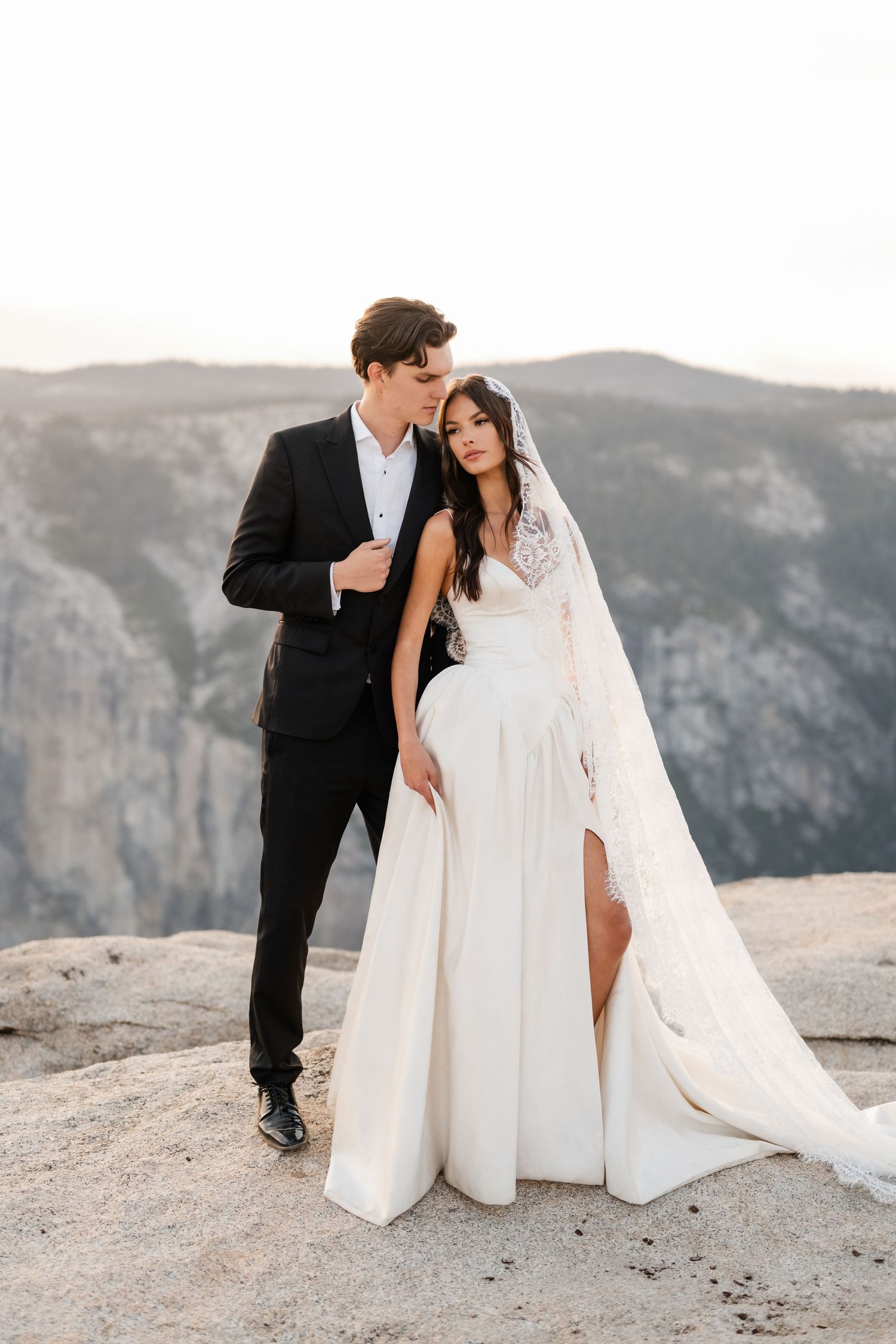 Bride and groom embrace on rocky mountain overlook, bride in white lace gown with veil, groom in black tuxedo.