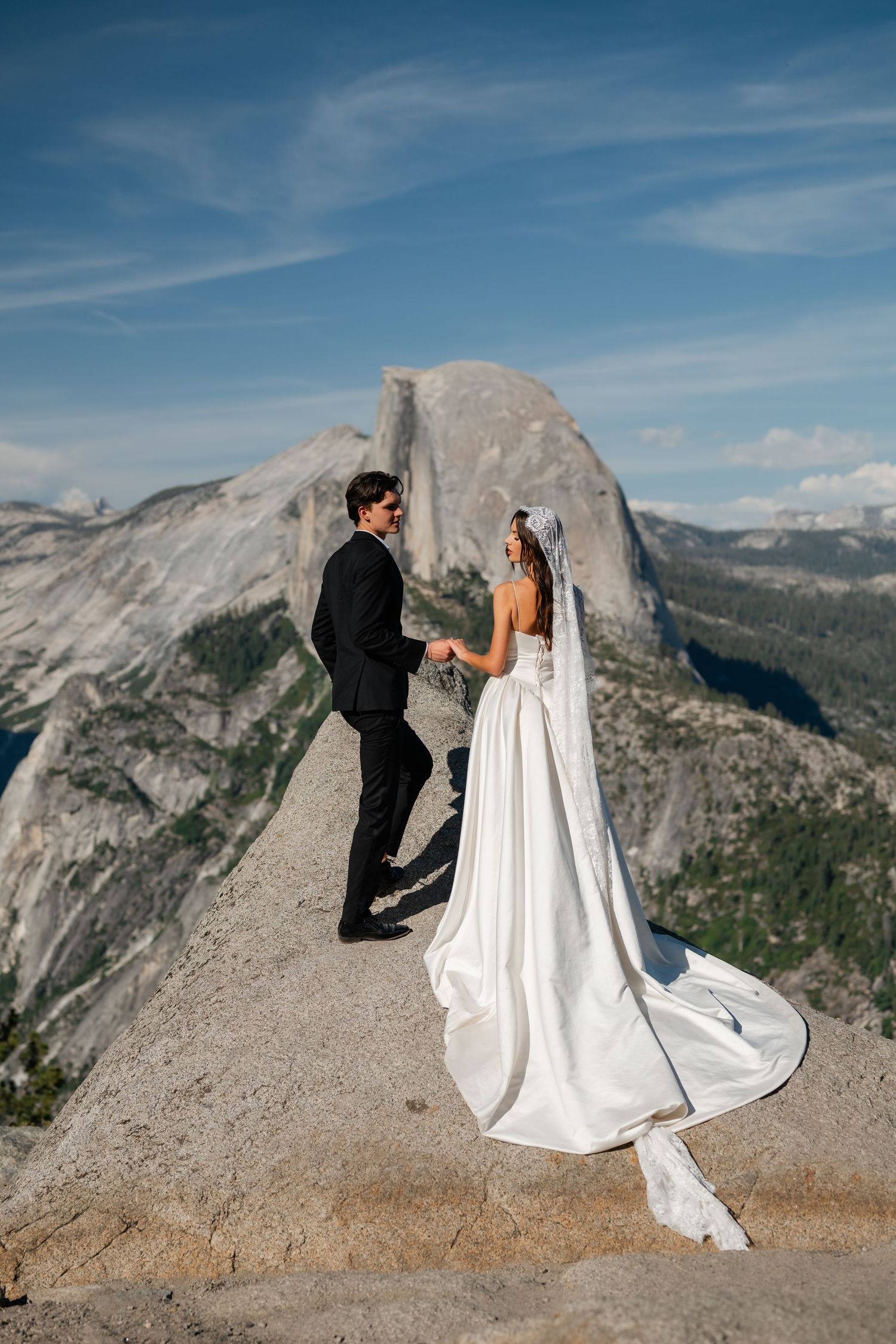 Bride and groom hold hands at Glacier Point, Yosemite, with Half Dome and mountain views behind them.